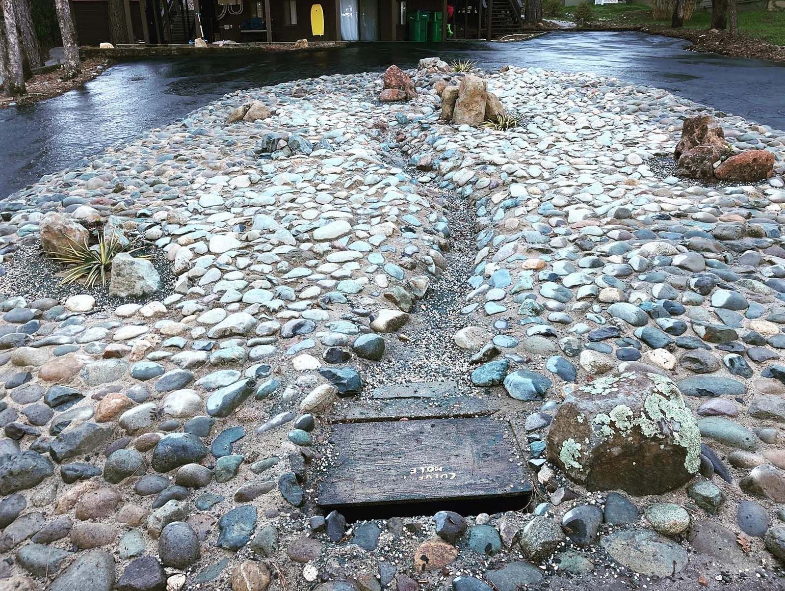 A dry creek bed made of various sized stones leading to a metal storm drain grate, surrounded by an asphalt driveway.