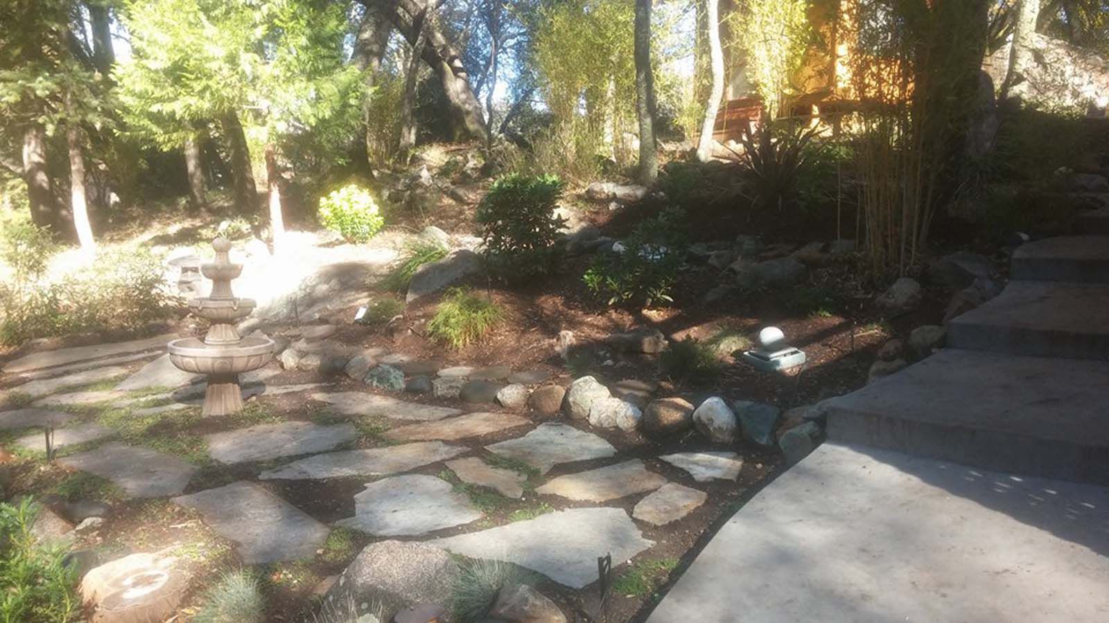 A stone pathway leads past a tiered fountain through a sunlit, landscaped garden with trees and shrubs.
