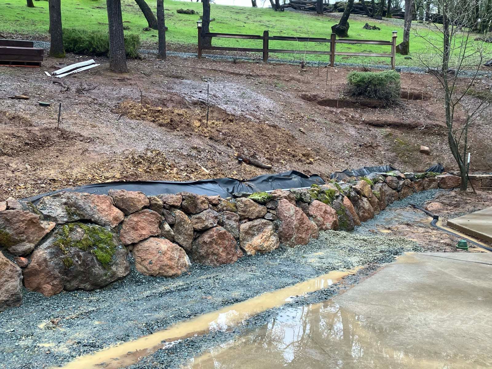A low stone retaining wall sits above a gravel drainage path with a large puddle near a concrete driveway.