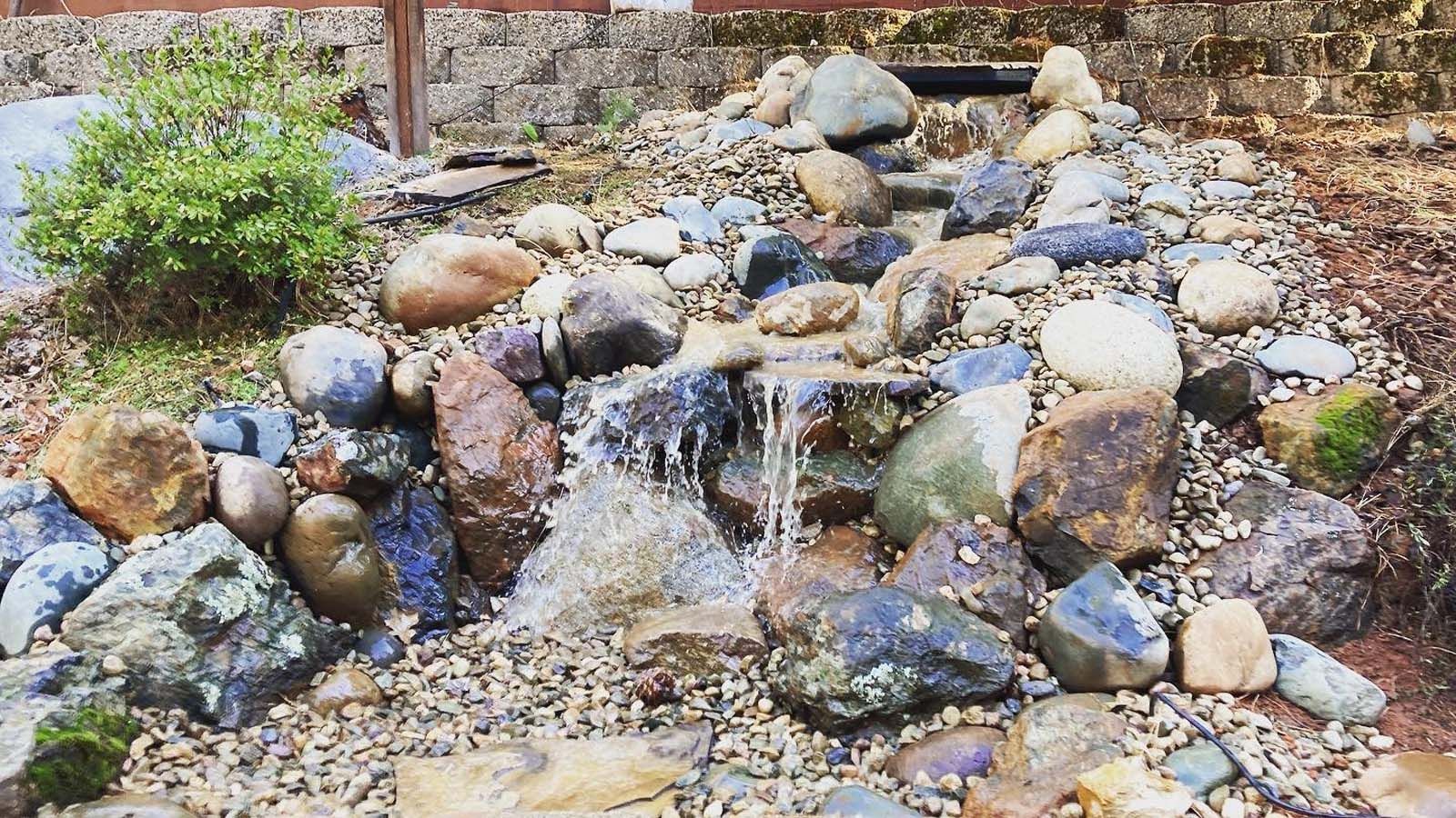 A small backyard water feature with cascading water flowing over decorative rocks and pebbles surrounded by landscaping.