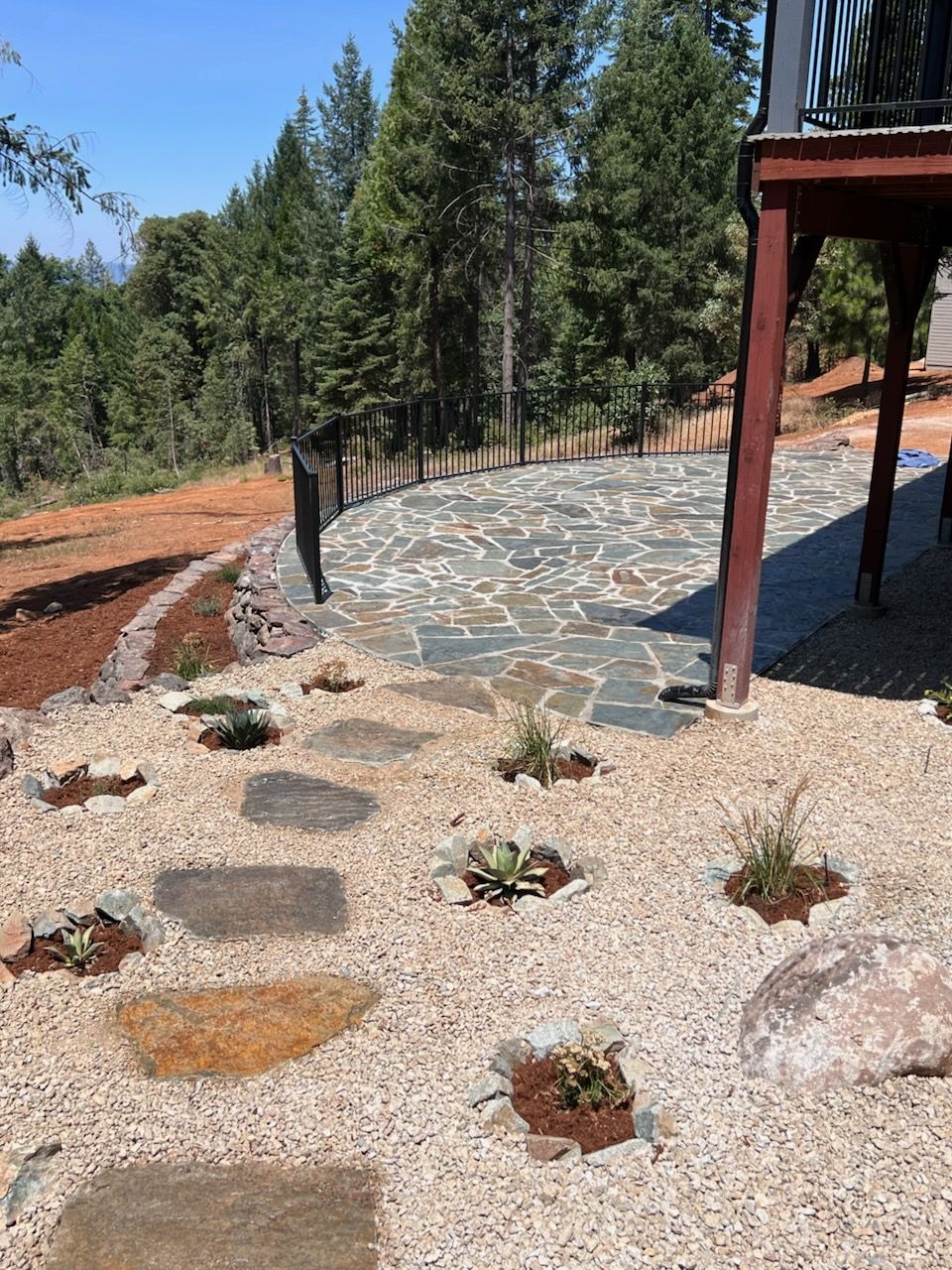 A stone path leads through a gravel garden with small plants toward a flagstone patio near a wooden deck and forest.