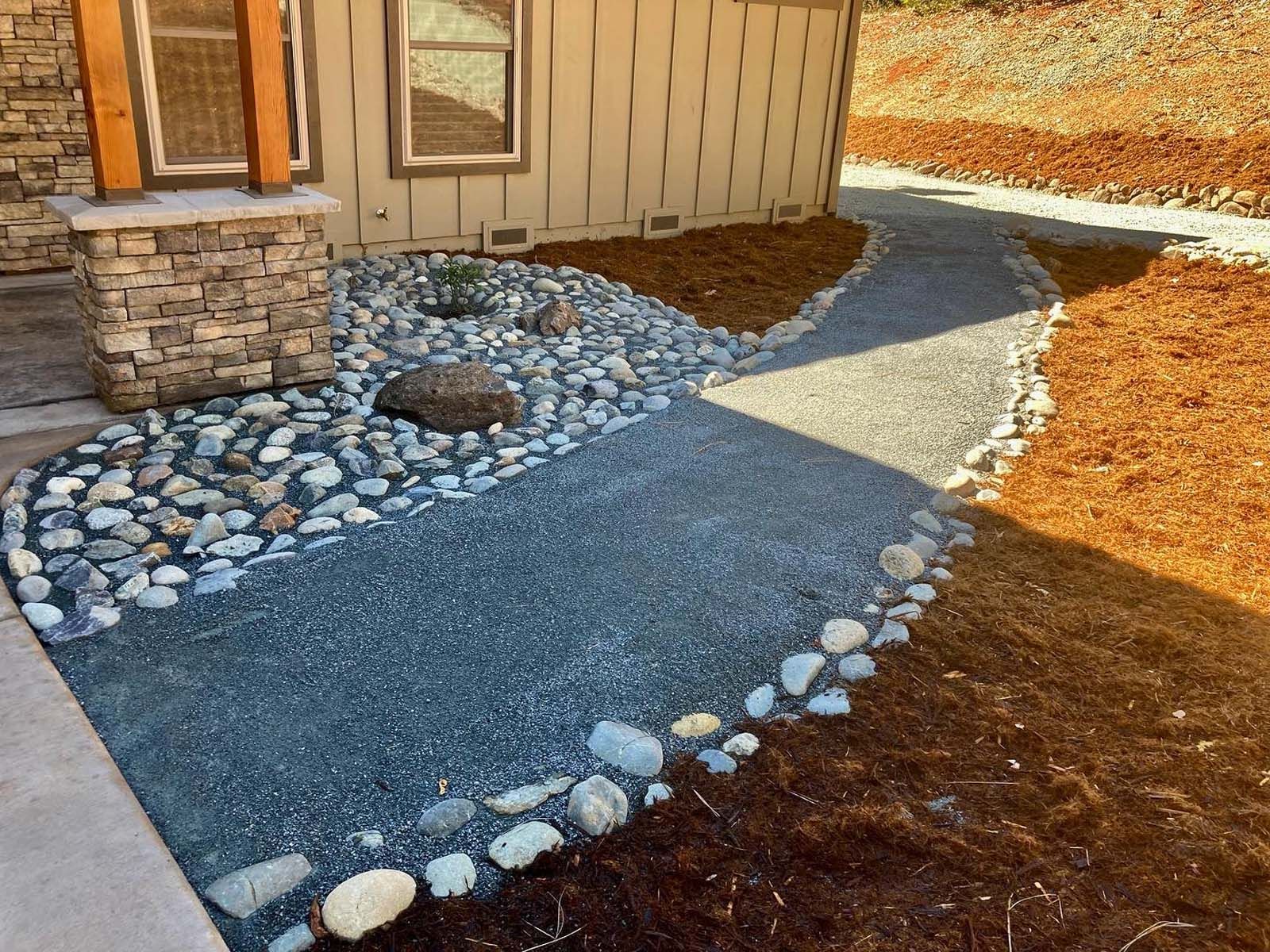 A stone-bordered gravel path winds through a landscape with mulch beds alongside a home exterior with stone accents.