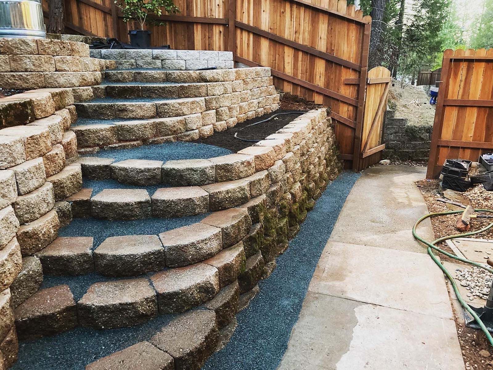 Stone stairs built into a backyard slope, leading up toward a wooden fence and gate, next to a concrete path.
