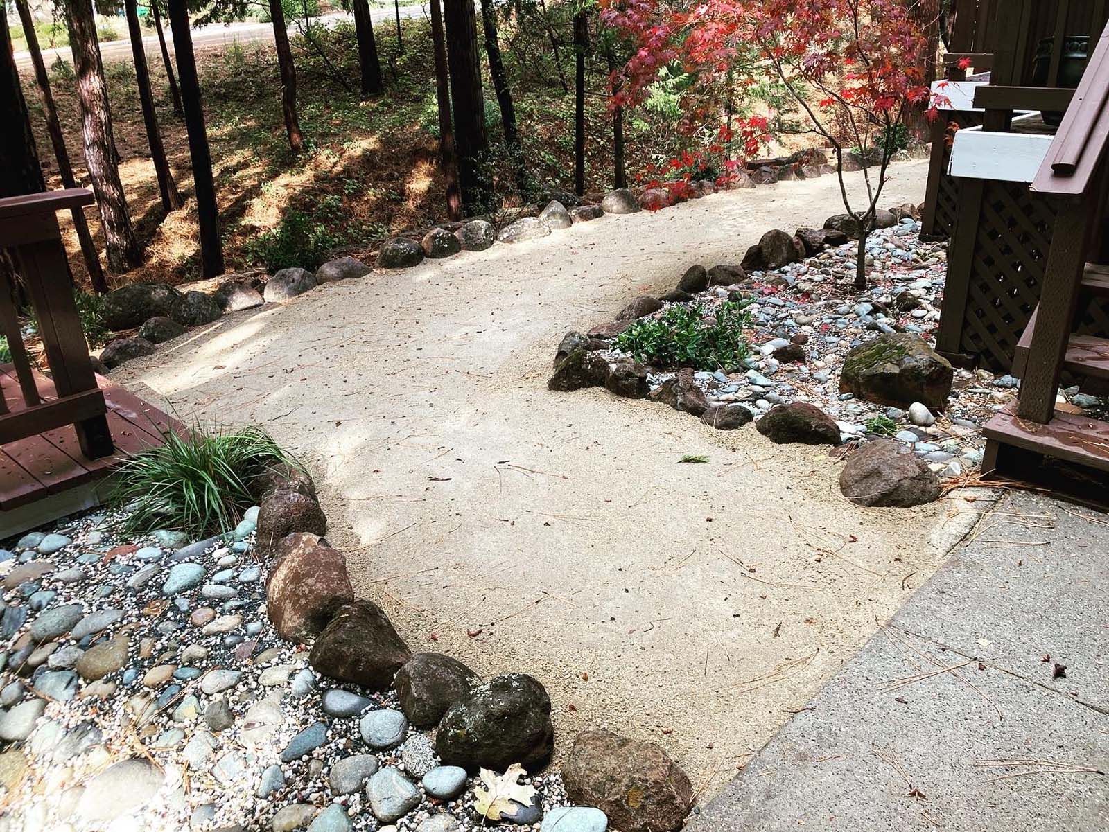 A gravel garden path curves between wooden deck stairs and a rocky landscaped bed in a wooded area.