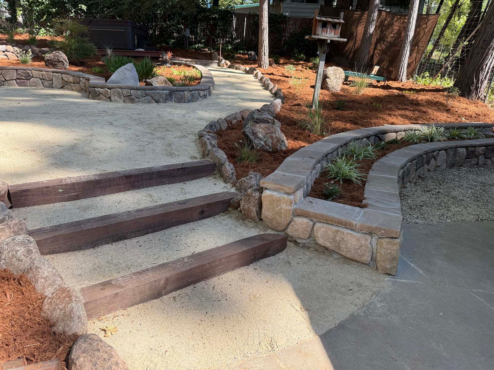A garden path with wooden stairs leads up to a stone retaining wall and landscaped area with mulch and a birdhouse.