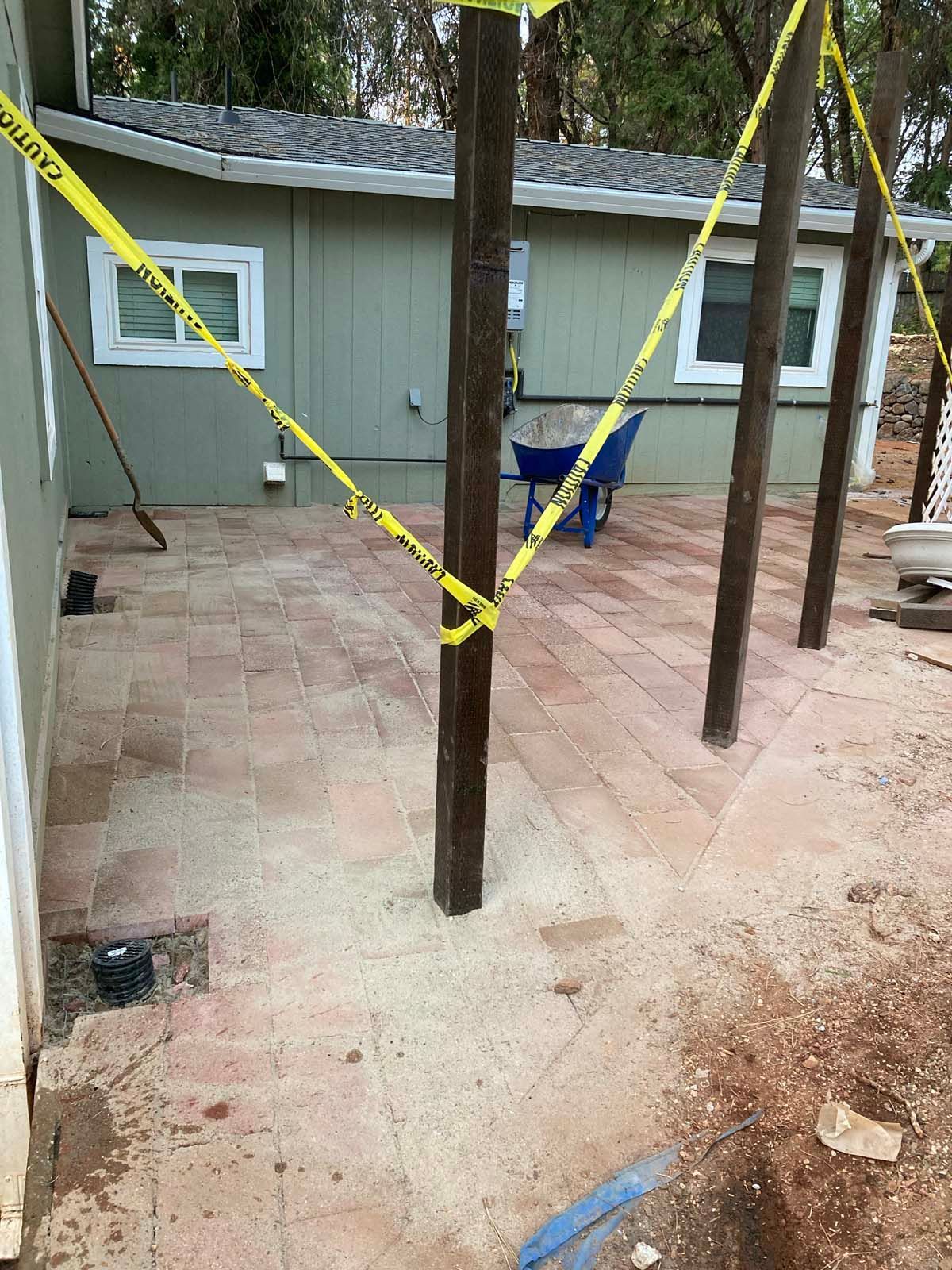 A recently installed light-brown brick patio sits in front of a gray house with two wooden support posts wrapped in tape.