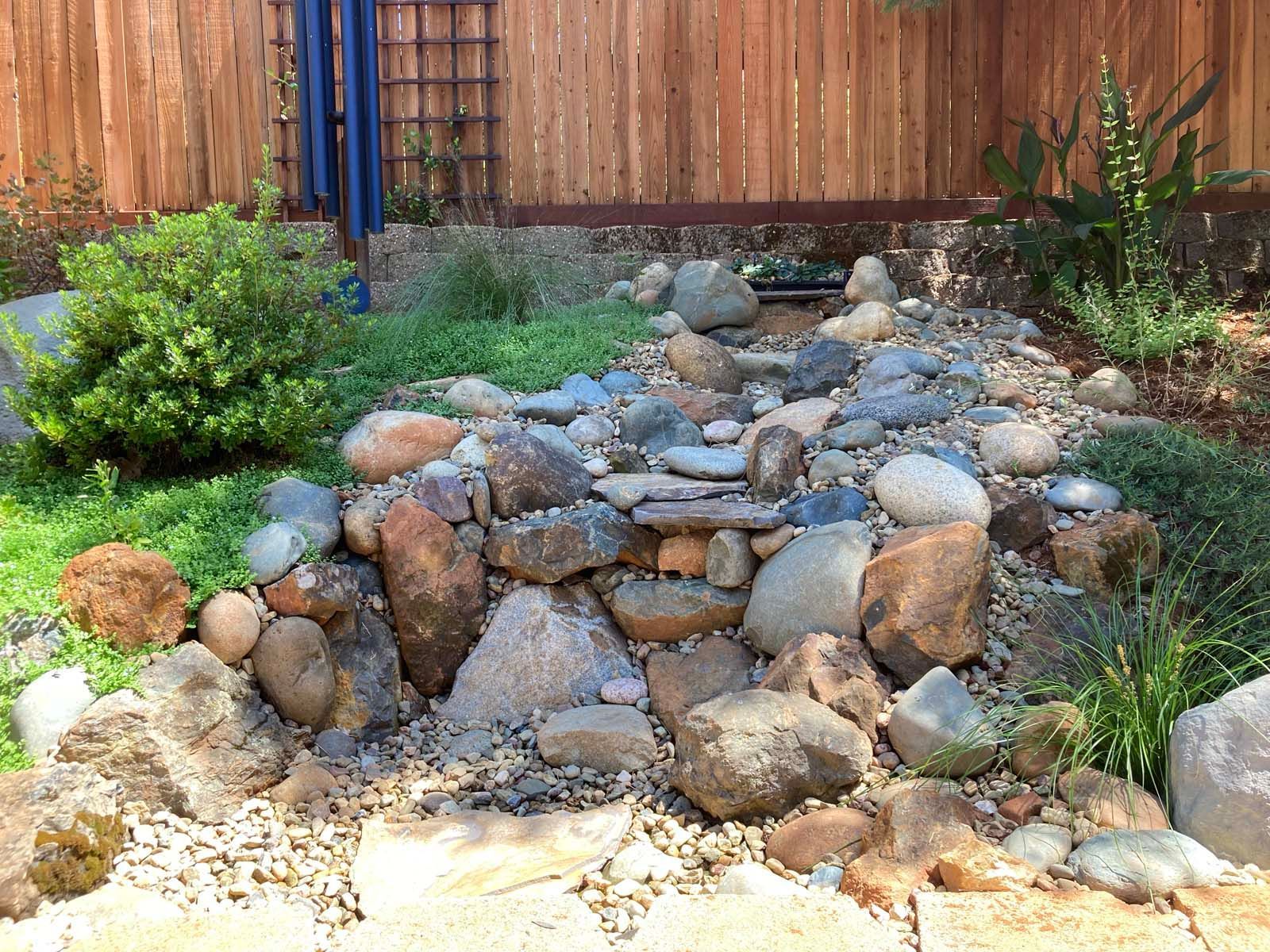 A rock garden feature with a cascading dry creek bed, bordered by lush green plants and a wooden fence backdrop.