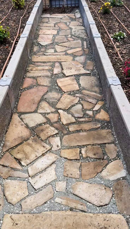 A stone flagstone pathway with gravel joints, enclosed by light gray concrete edging in a garden setting.