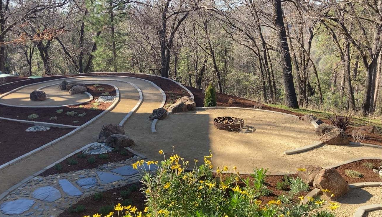 A landscaped garden featuring a winding stone path, gravel sections, scattered rocks, and small plants against a forest.