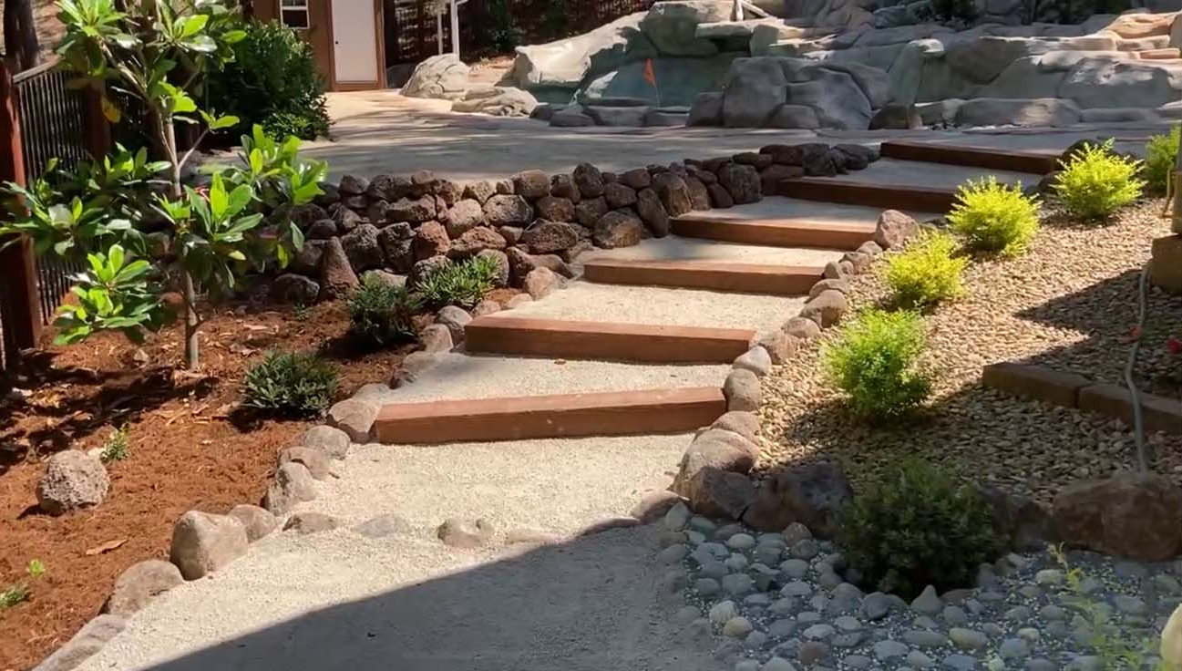 A landscaped outdoor path featuring tiered wooden steps, gravel, and small green shrubs leading toward a rock garden area.