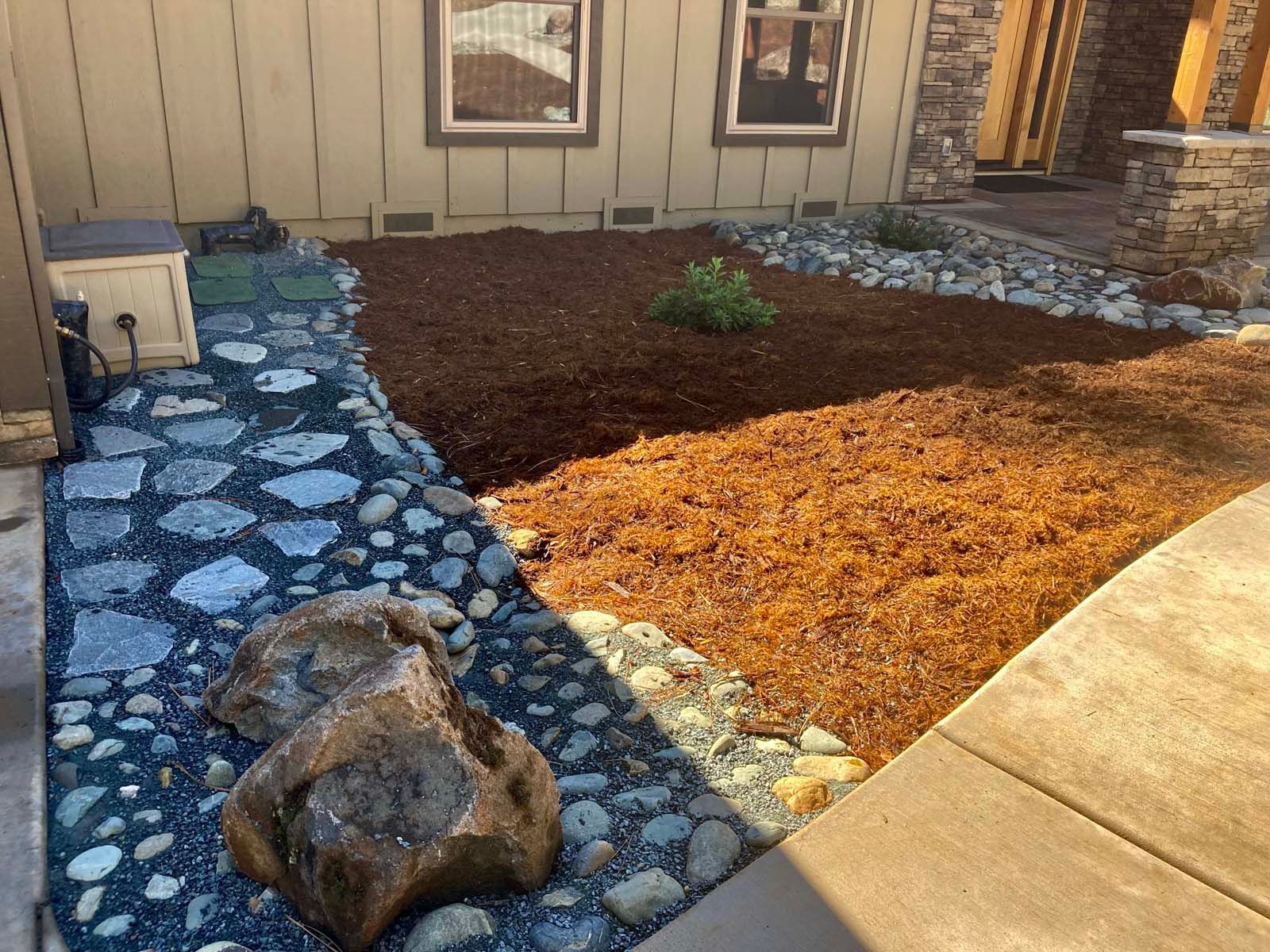 A stone walkway borders a yard with brown mulch and a small green shrub in front of a house with two windows.