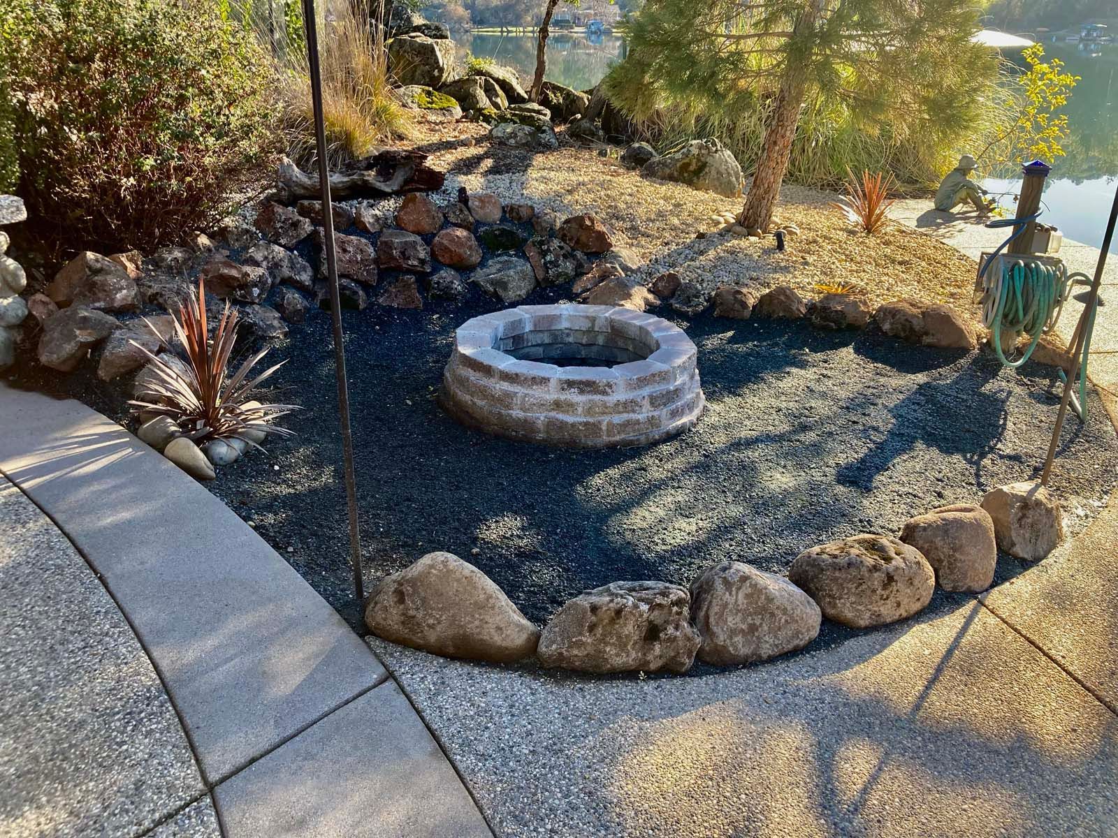 A circular fire pit made of light-colored bricks sits in a landscape bed filled with dark gravel, bordered by large rocks.