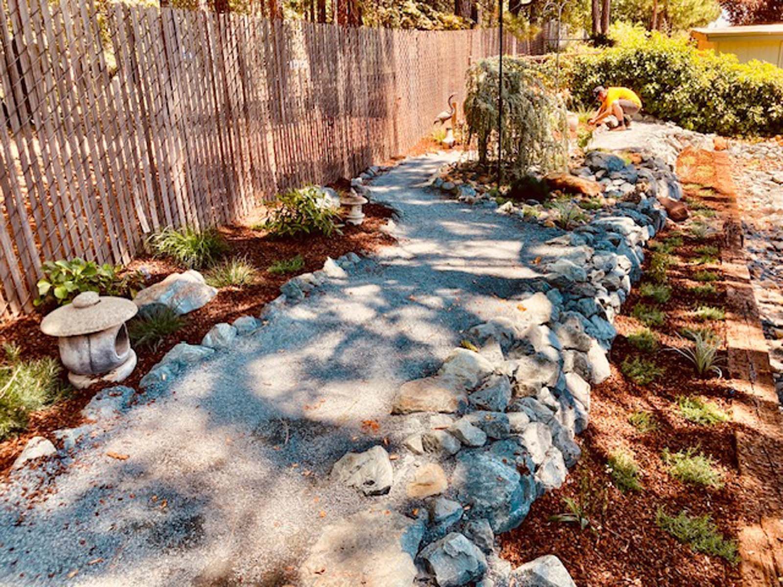 A winding gravel path lined with stones through a garden, with a person gardening in the background near a wood fence.