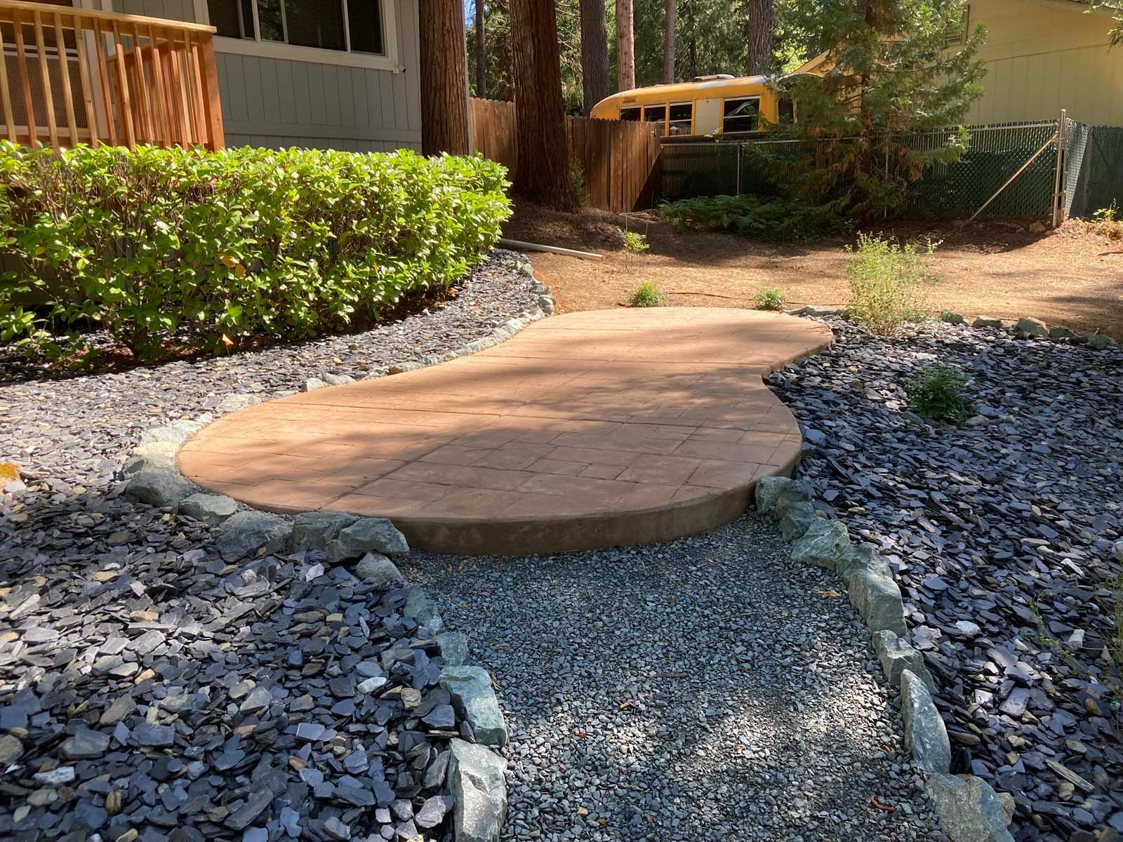 A raised, kidney-shaped wooden platform set within a gravel path surrounded by dark landscaping stones and green bushes.