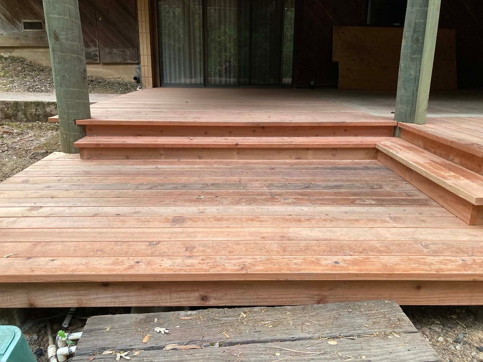 A newly built wooden deck featuring two levels connected by stairs, leading toward a glass sliding door entrance.