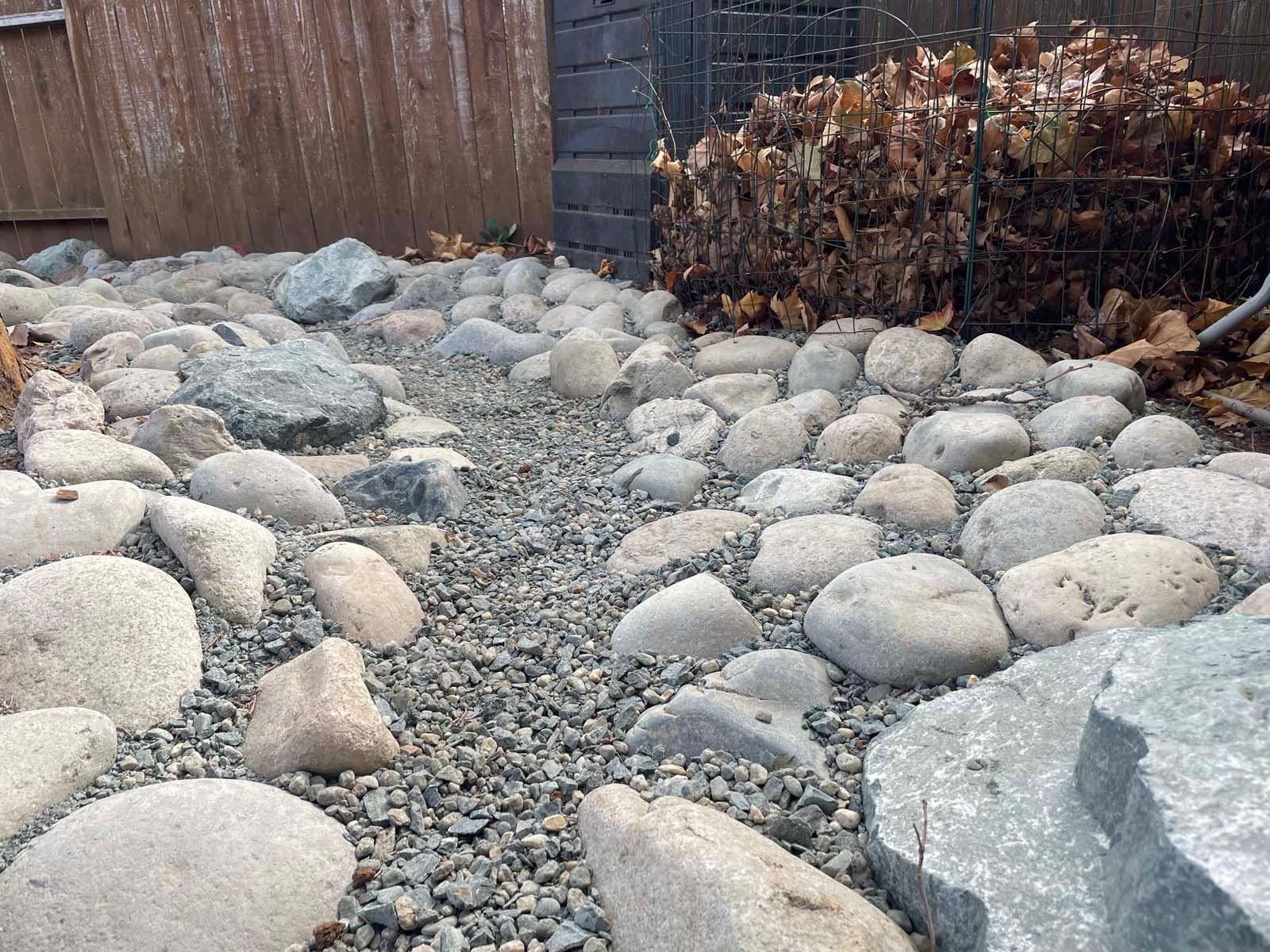 A dry creek bed landscaping feature with grey river rocks, small gravel, a wooden fence, and a compost bin of dead leaves.
