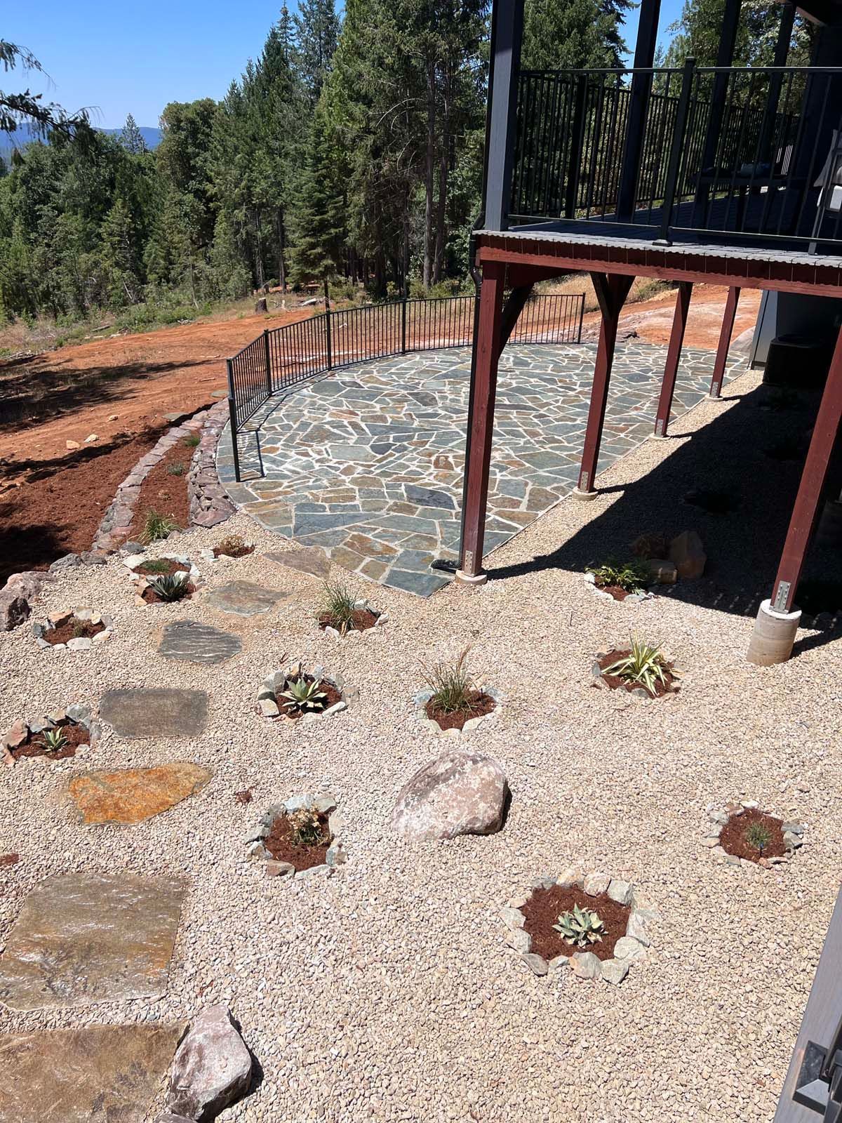 A stone patio with a wood railing under a deck, overlooking a tree-lined hillside with a gravel path and small plantings.