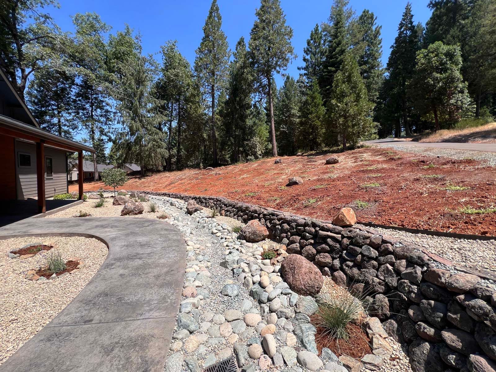 A stone retaining wall curves beside a dry creek bed and a paved walkway in a pine forest setting with a gravel slope.