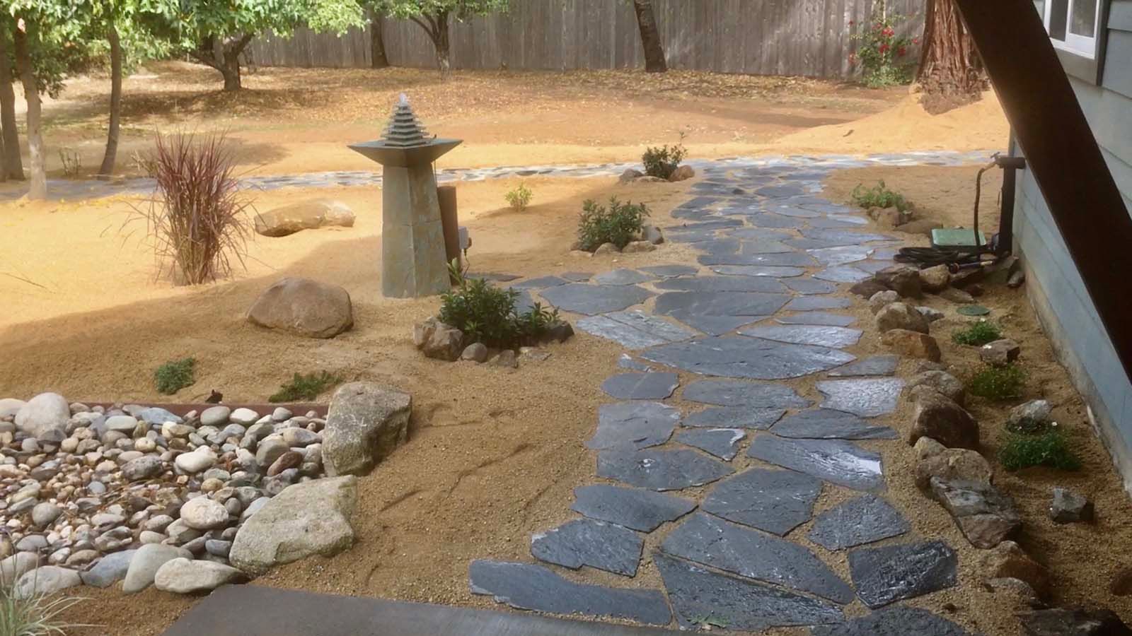 A stone flagstone walkway leads through a landscaped yard with tan gravel, rocks, a birdbath, and scattered shrubs.