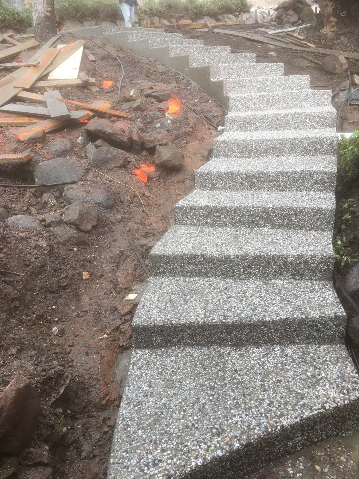 A curved outdoor staircase made of grey speckled concrete leads up a dirt slope, with construction materials to the left.