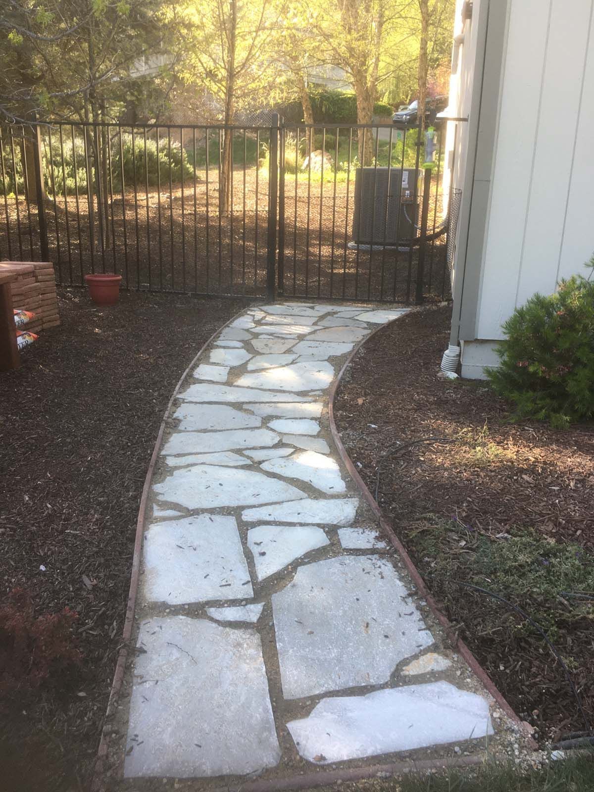 A flagstone path winds toward a metal gate beside a house, surrounded by dark mulch and trees.