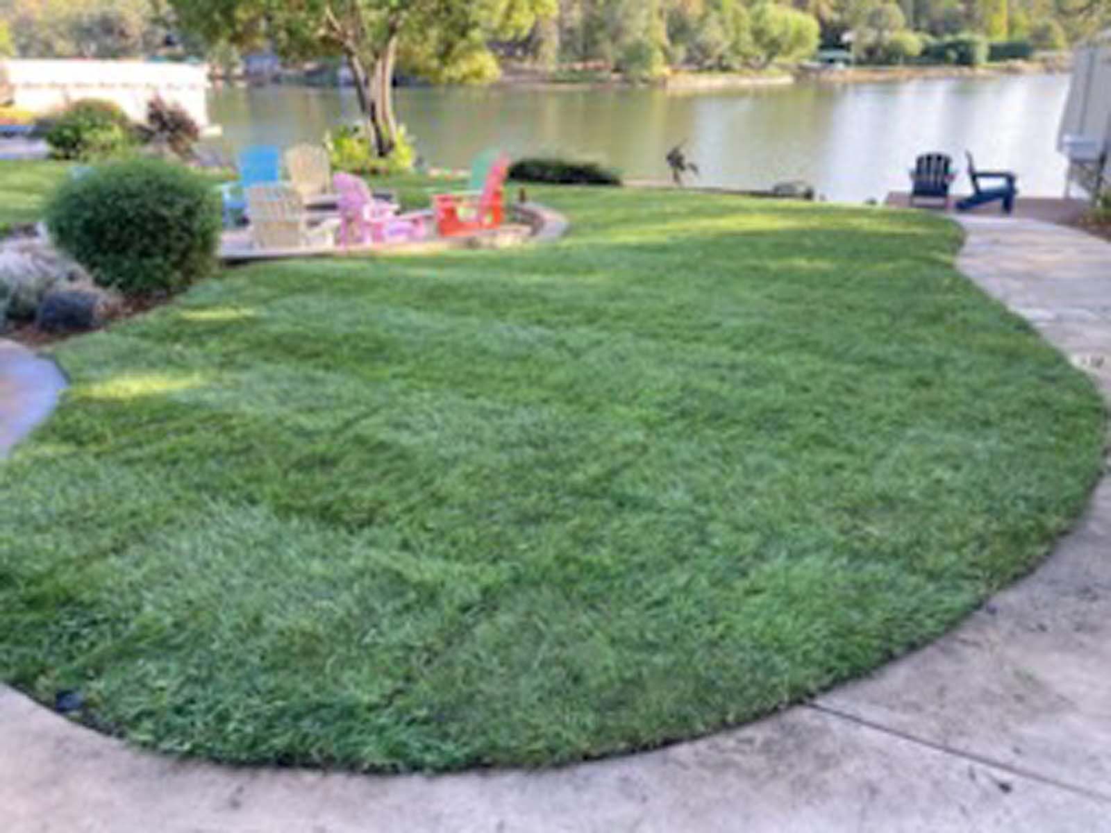 A lush green lawn curves around a stone patio with colorful chairs overlooking a calm lake on a sunny day.