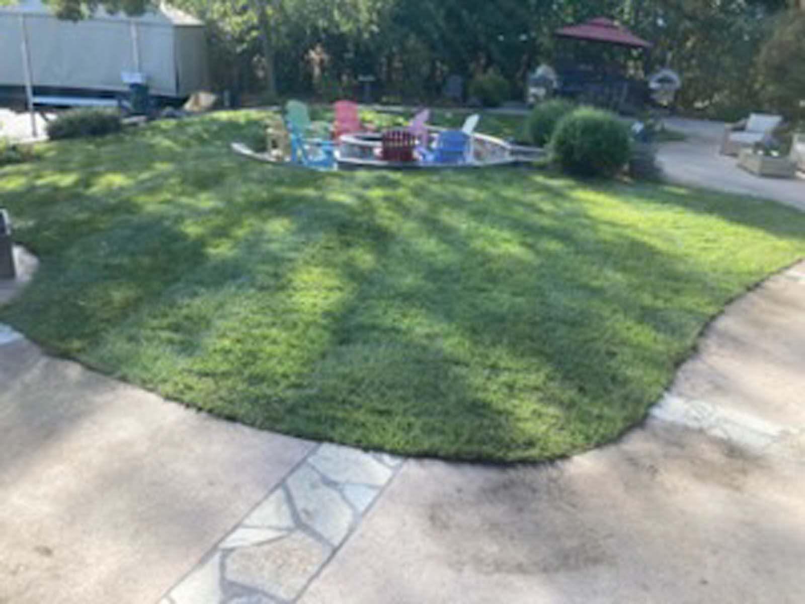 A grassy backyard with a curved stone fire pit area featuring several colorful chairs, surrounded by a concrete patio.