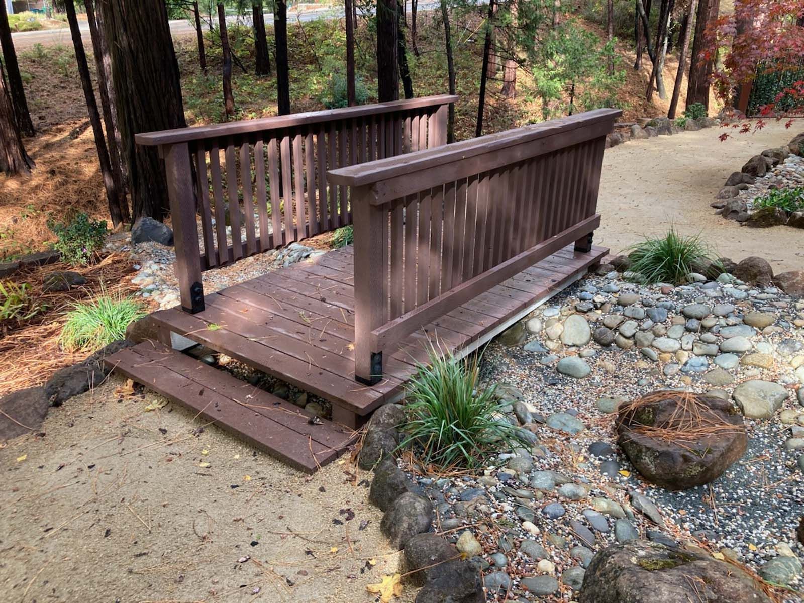 A brown wooden footbridge spans a dry, rock-filled creek bed in a forested setting with a tan dirt path.
