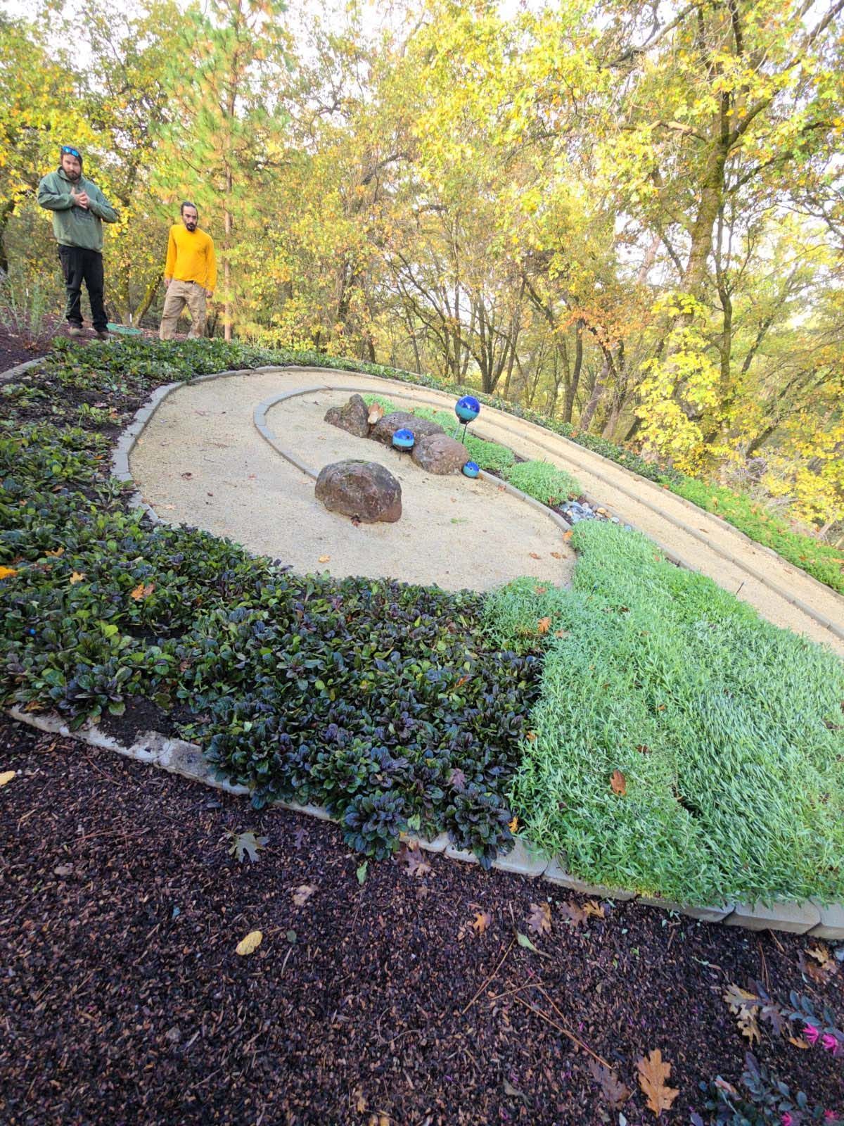 Two people stand above a manicured, spiral garden path with central rocks, surrounded by trees in a park setting.