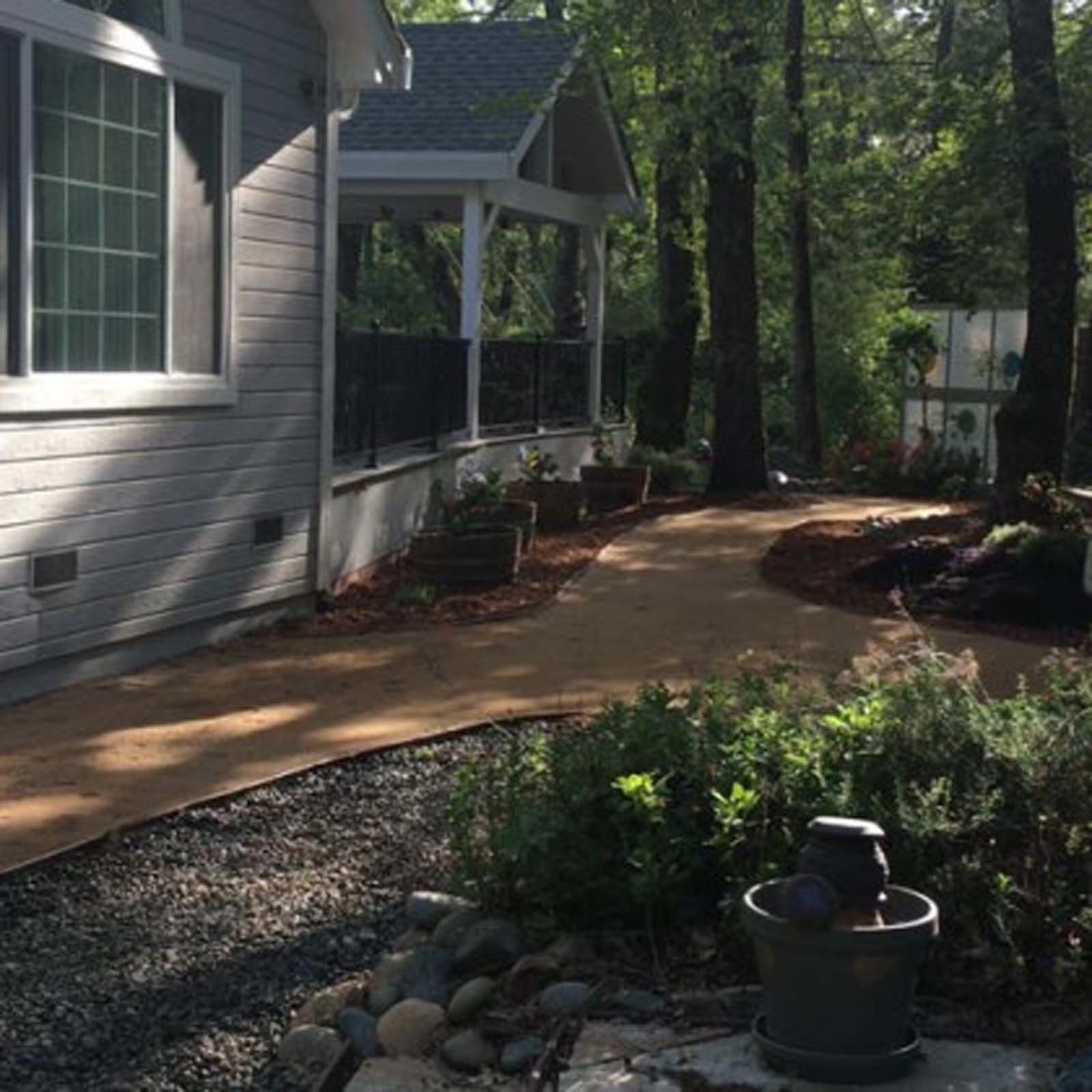 A house with gray siding and a porch overlooks a winding dirt path through a wooded yard with gravel and small plantings.
