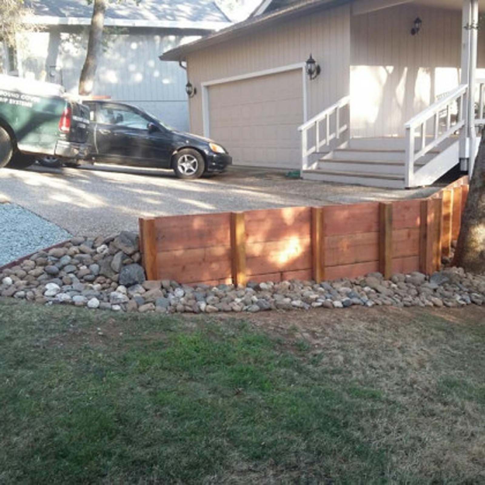A new wooden retaining wall with a stone base borders a driveway next to a house with a staircase.