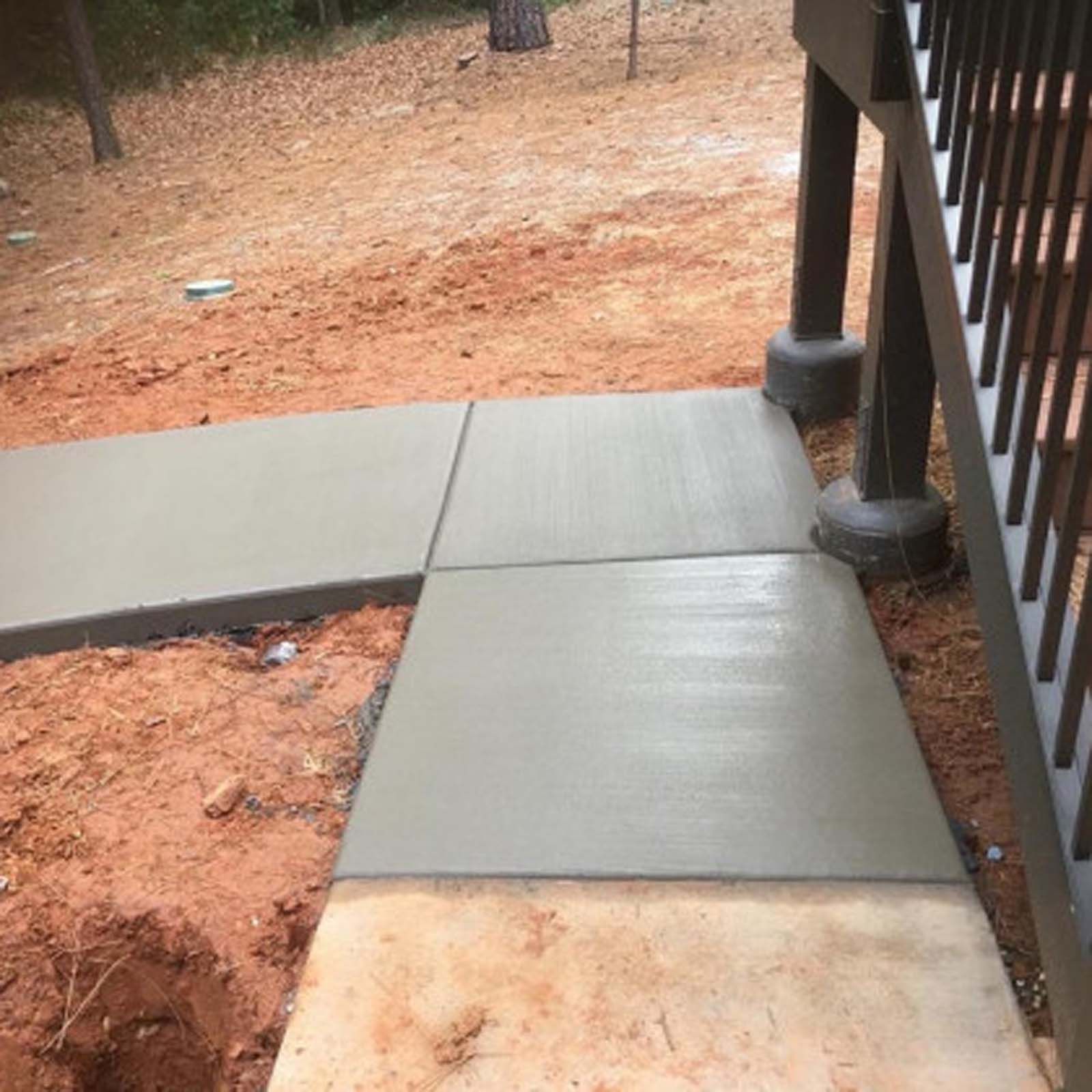 Freshly poured concrete sidewalk sections installed next to a dark wood deck support post and red dirt yard.