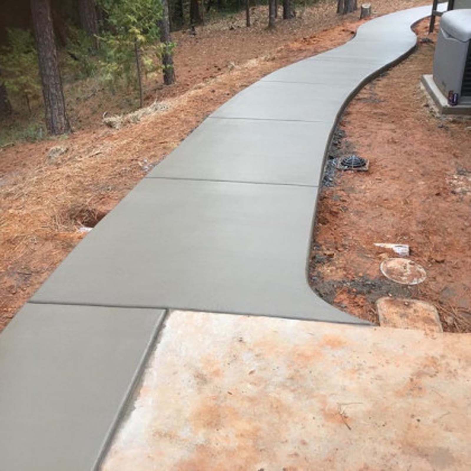 A new, curved gray concrete sidewalk winds through a wooded residential area, connecting to a concrete slab.