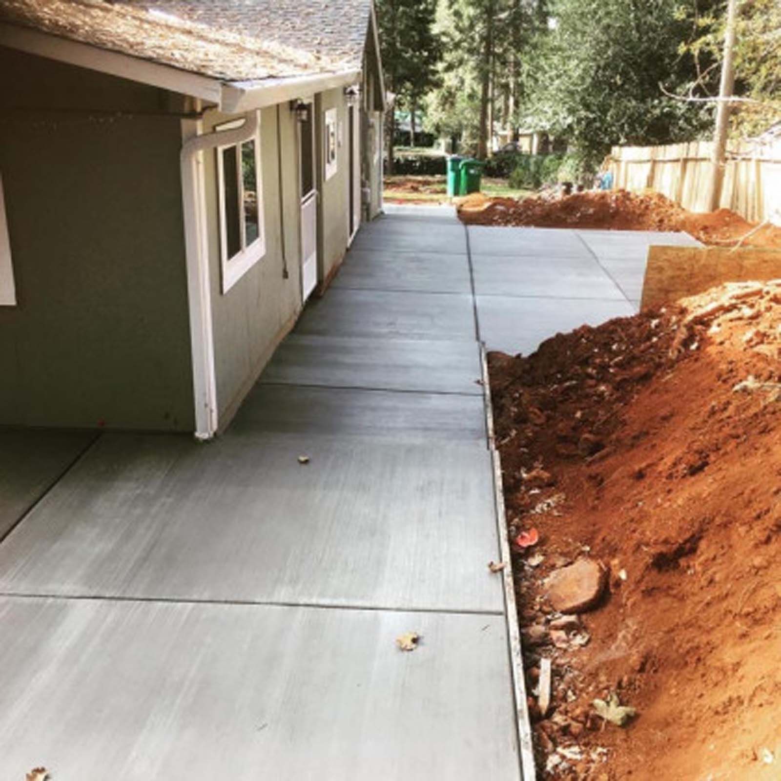 A new concrete walkway alongside a house, next to an area of excavated red dirt and unfinished landscaping.