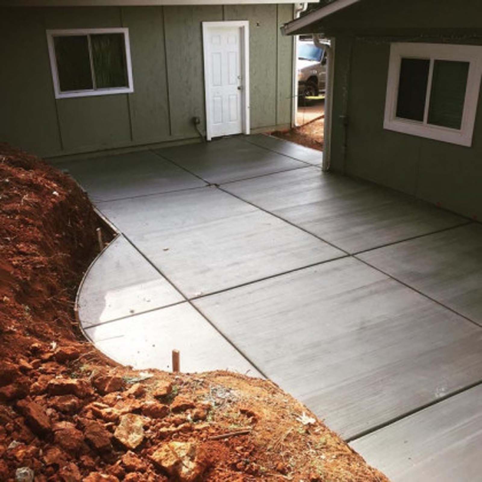 New concrete driveway and patio area next to a green house, with an unfinished dirt slope on the left.