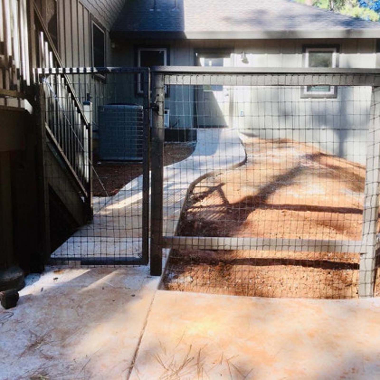 A black metal gate and wire mesh fence installed on a concrete patio next to a house staircase.