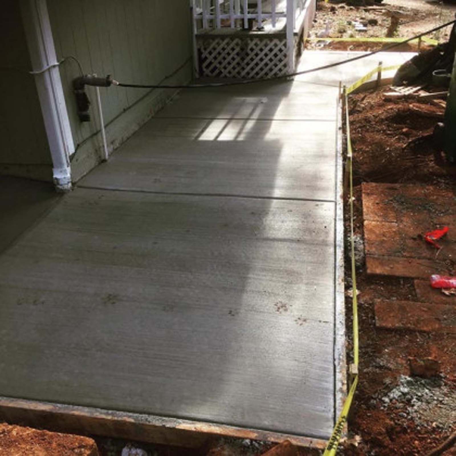 Newly poured concrete walkway bordering a house, framed by wooden forms and surrounded by dirt.