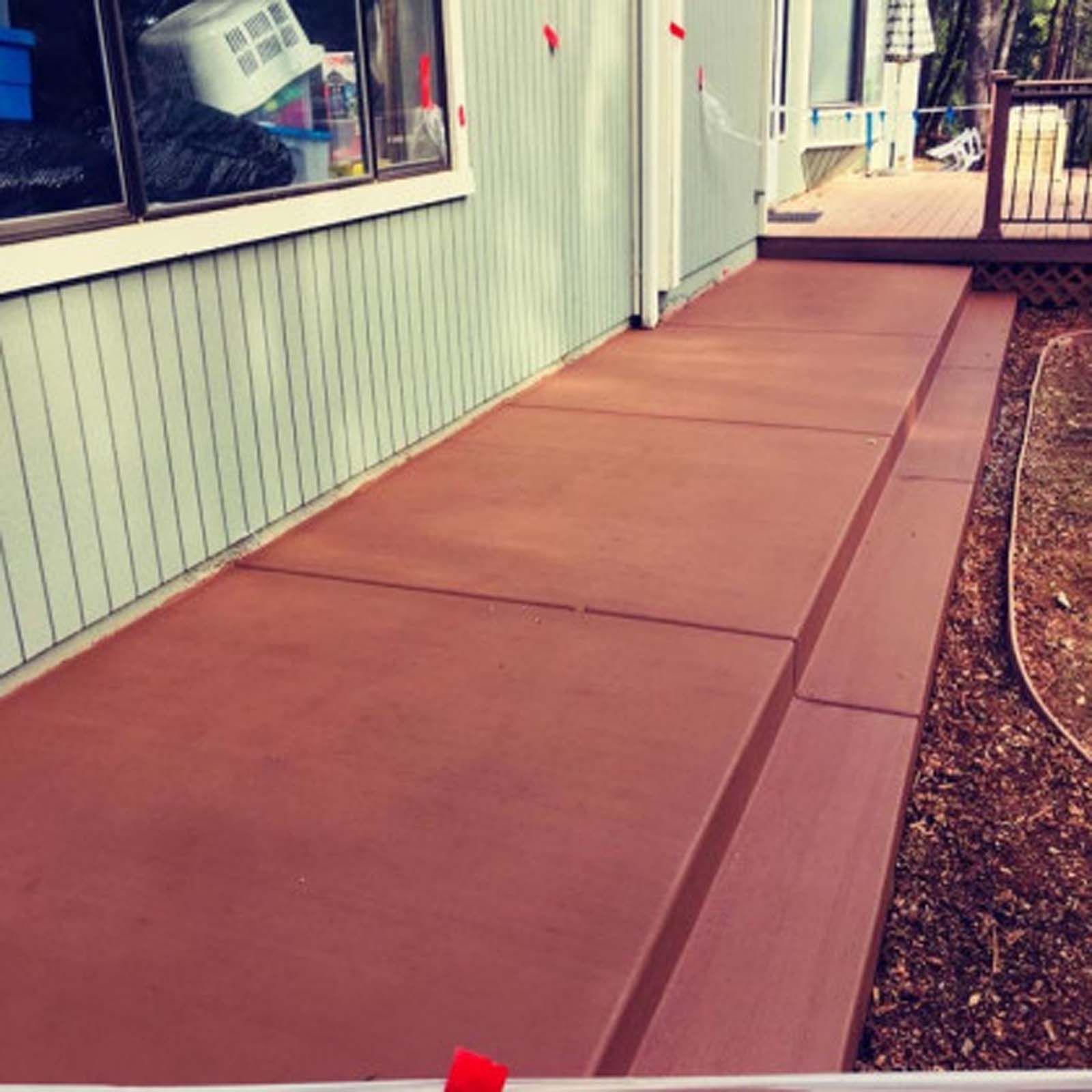 A freshly poured, terracotta-colored concrete walkway runs along the side of a house with light green siding.