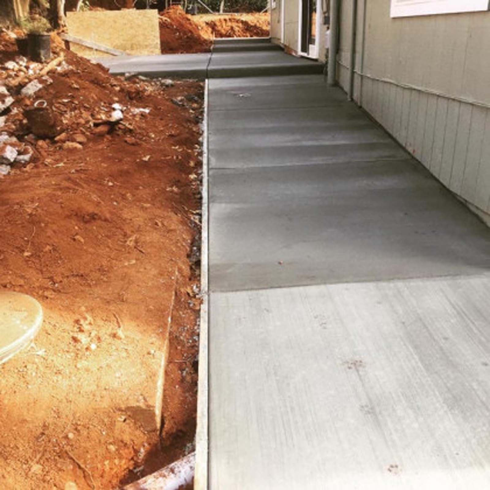 Newly poured concrete walkway running alongside a residential building next to a dirt embankment.