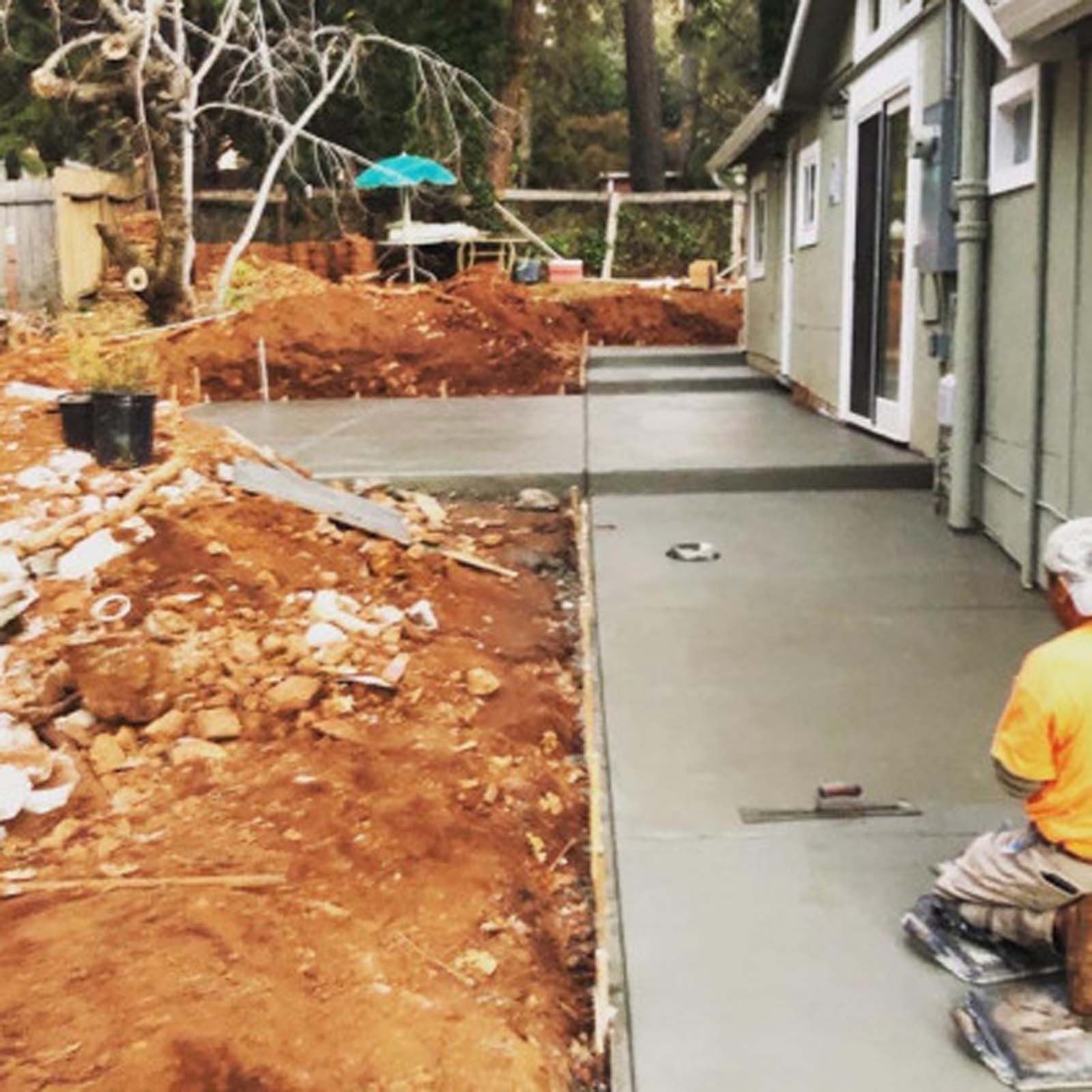 A worker in a yellow shirt finishes a new concrete patio extending from a house alongside a pile of excavated red dirt.