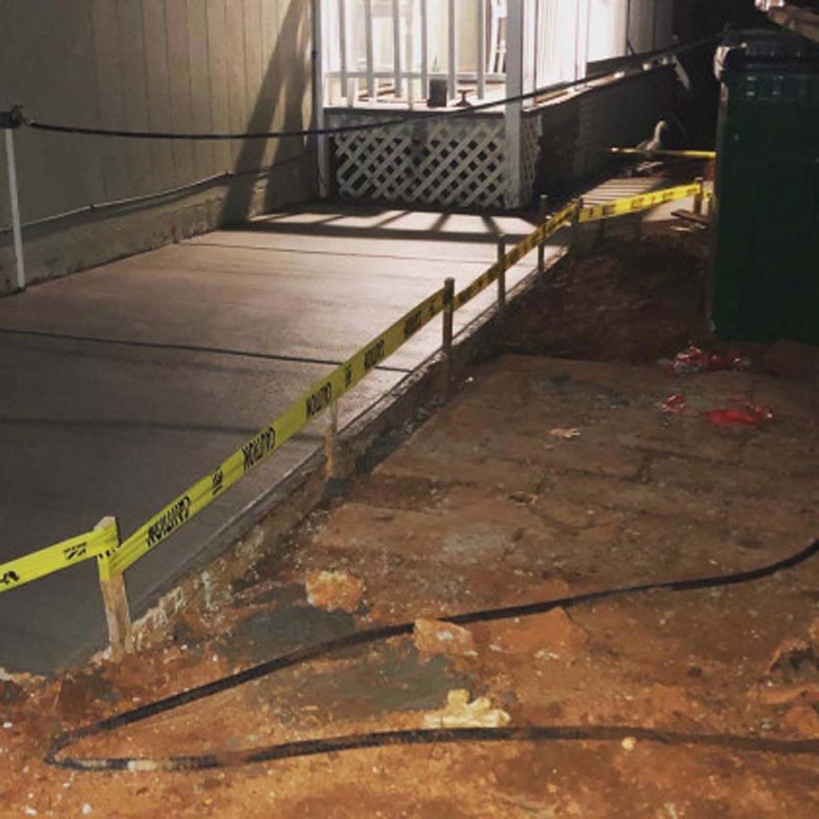 A sidewalk under construction at night, cordoned off with yellow caution tape, with a green trash bin nearby.