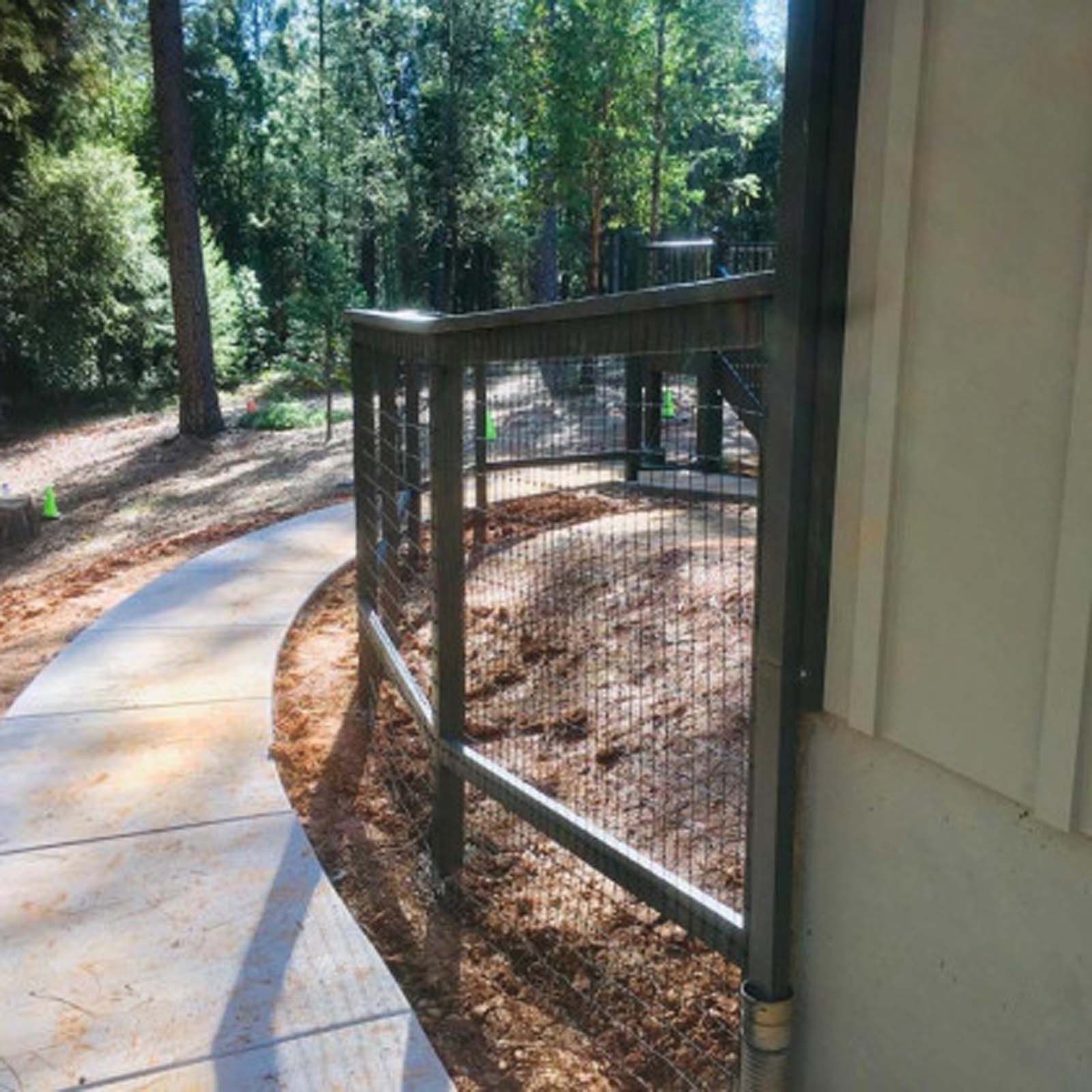 A curved, dark metal railing with wire mesh panels lines a concrete walkway next to a building, overlooking a wooded area.