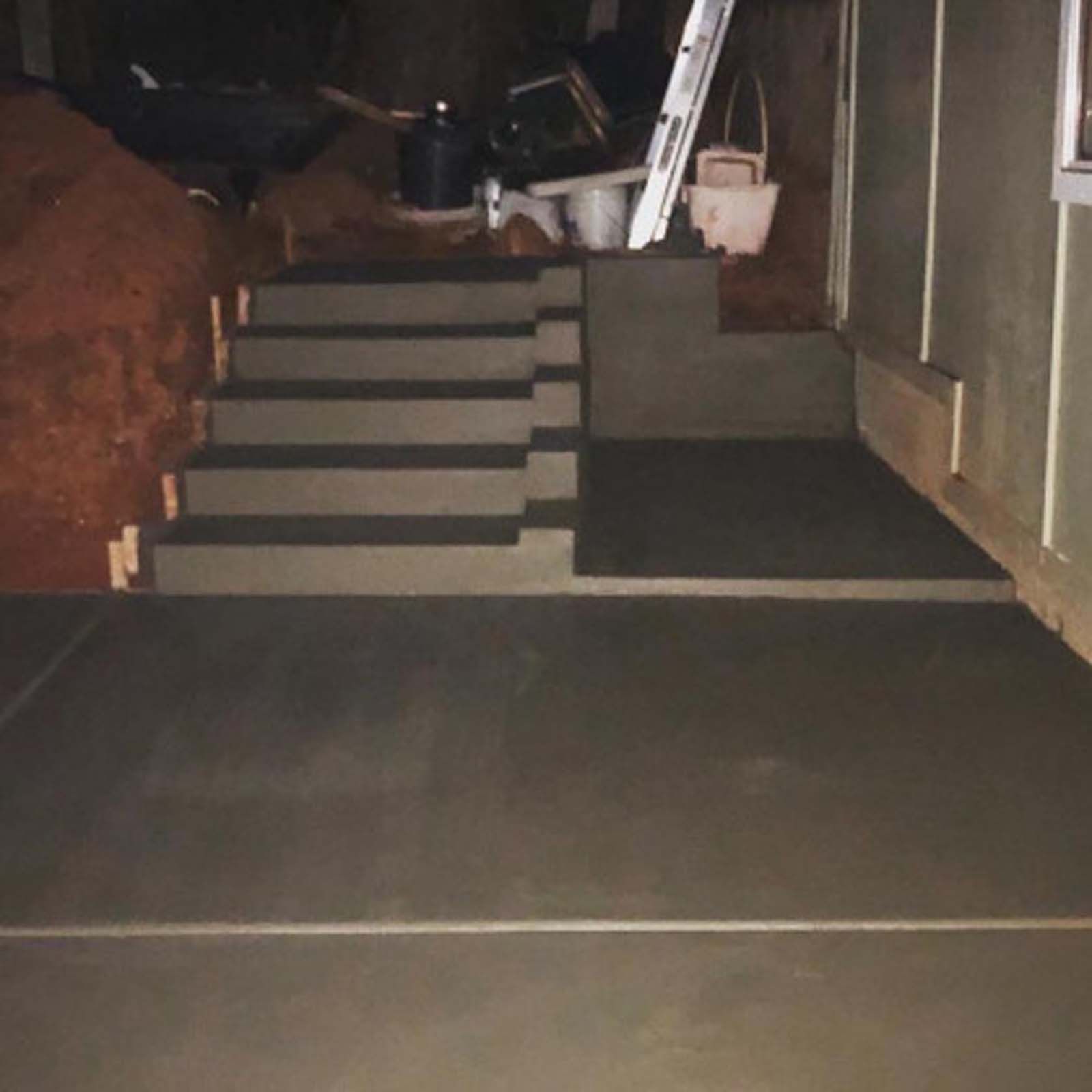 Freshly poured concrete stairs and a landing alongside a dirt embankment and the side of a building at night.
