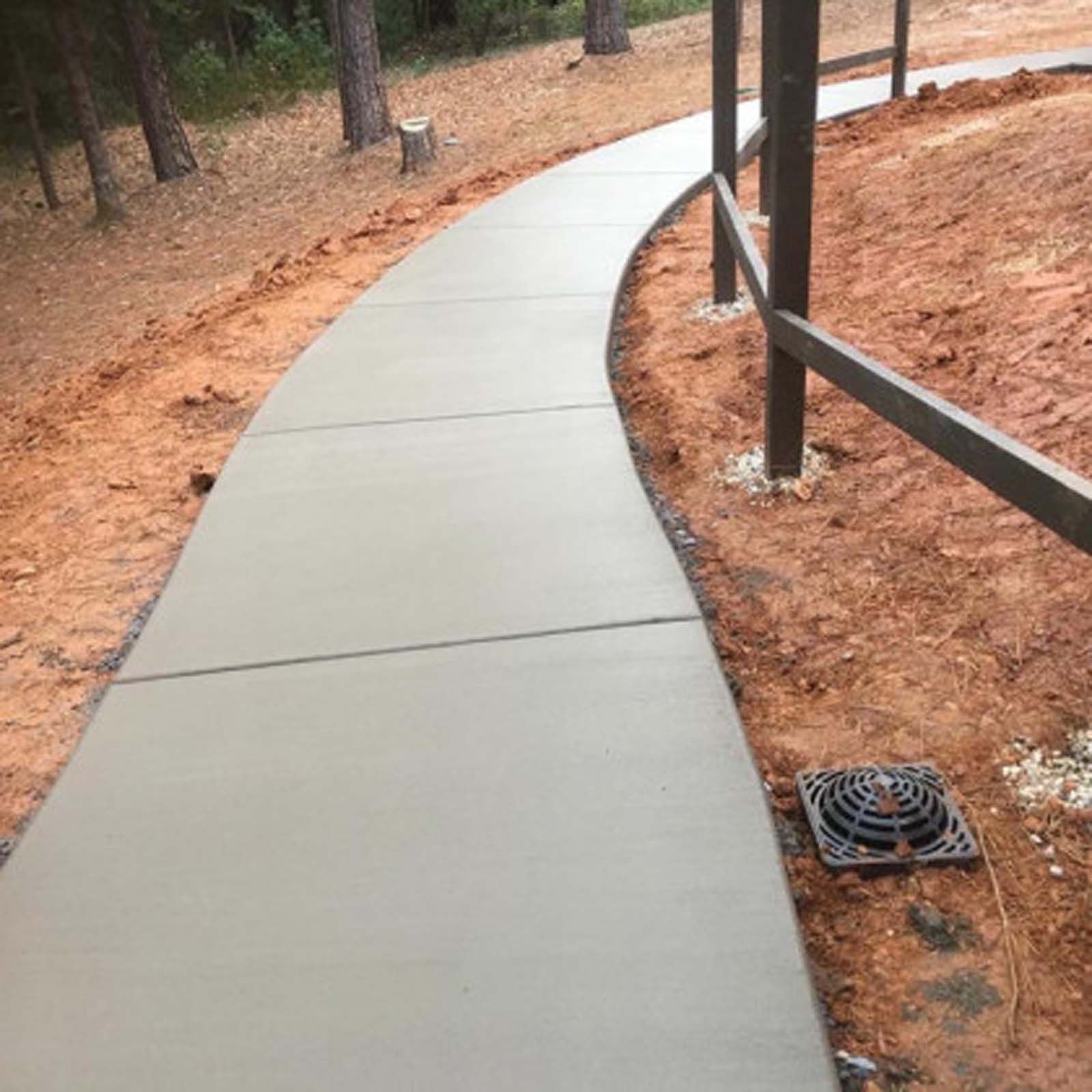 A newly poured concrete walkway curves through a wooded area next to a wooden post fence and a drainage grate.