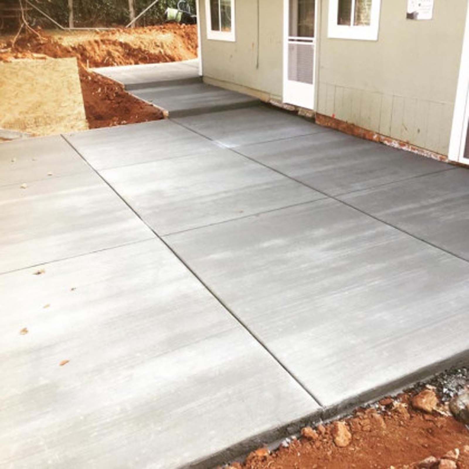 A newly poured concrete patio outside the side entrance of a building, surrounded by dirt and landscaping.