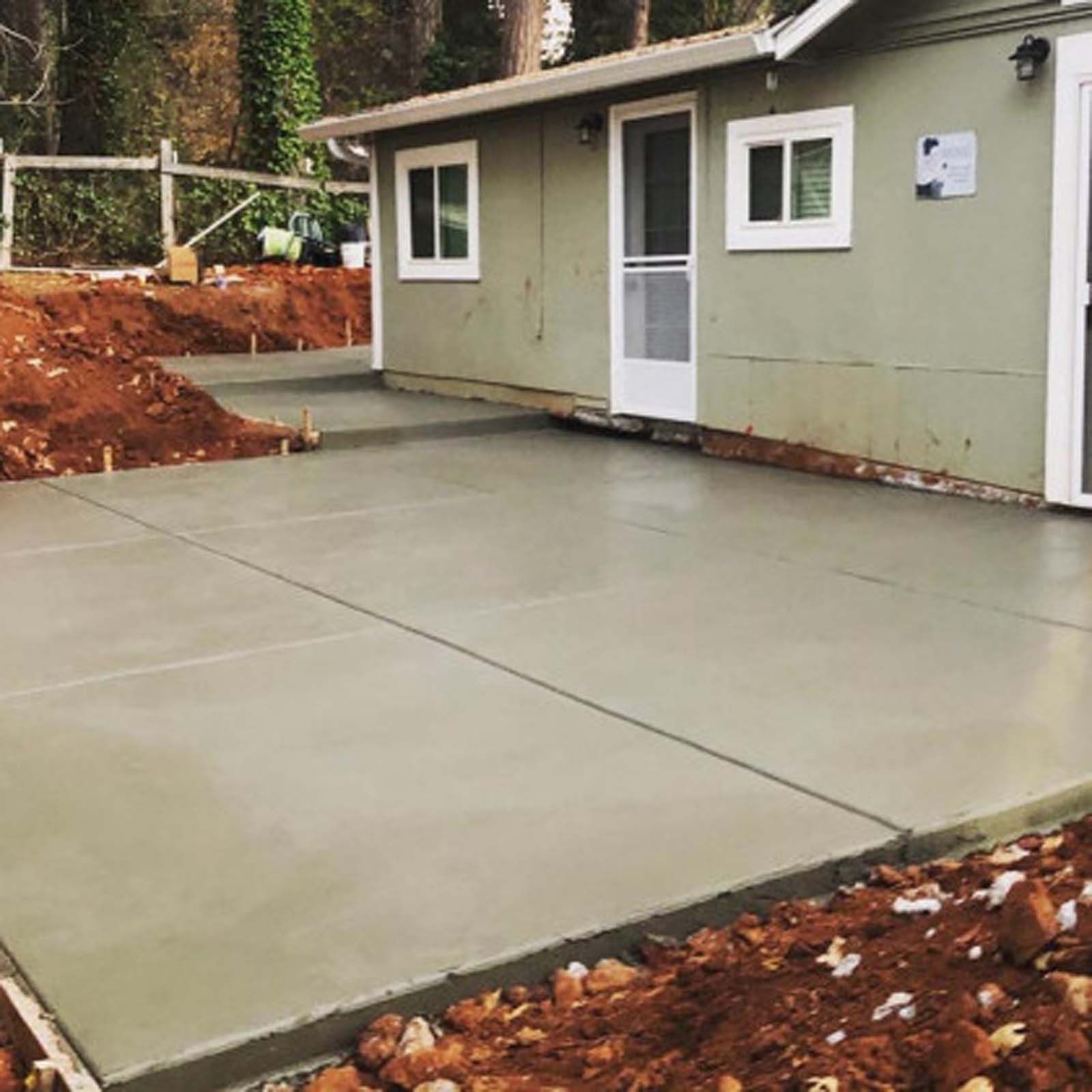 A light green house with a newly poured, smooth concrete patio in the foreground, surrounded by fresh dirt and landscaping.