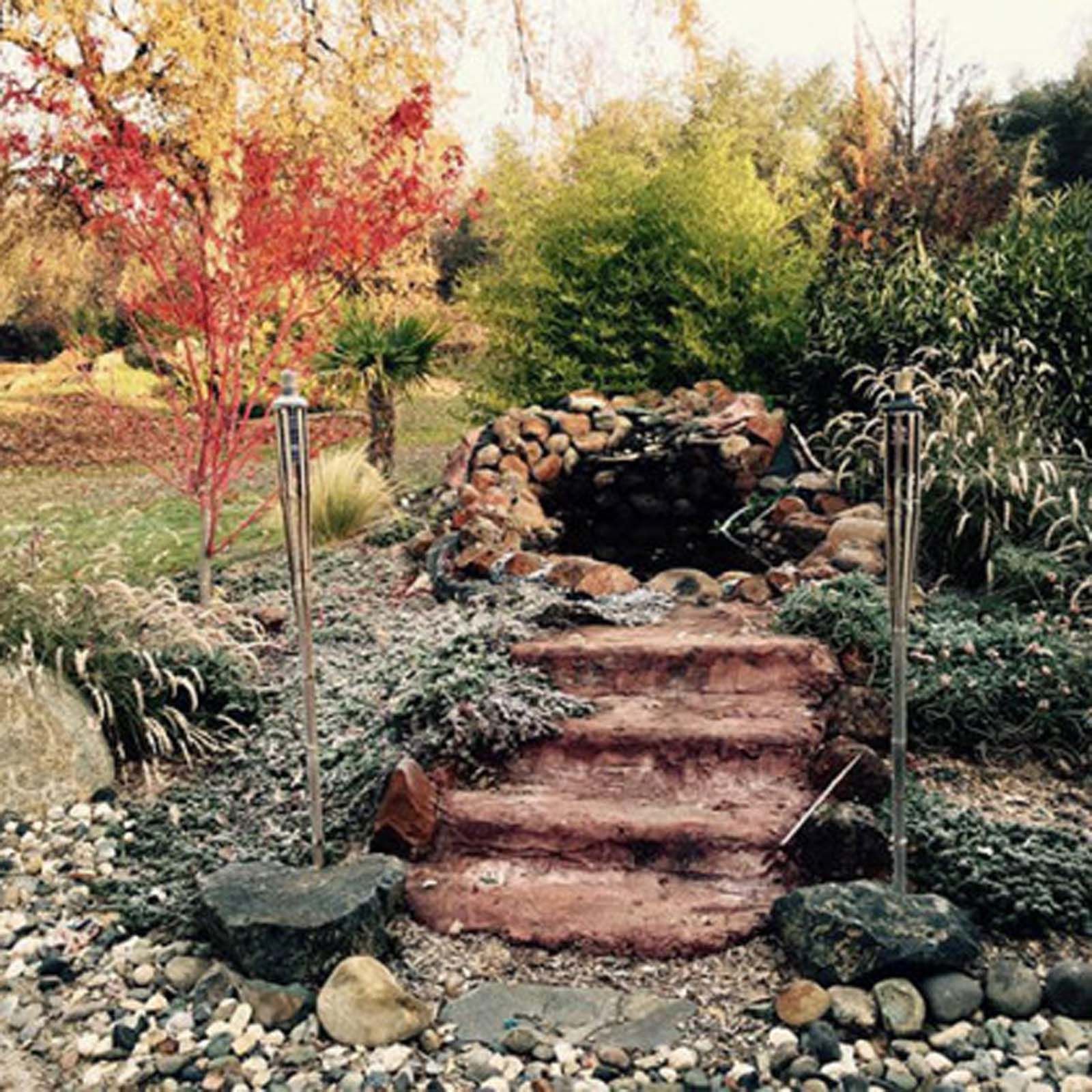 Stone stairs lead up to a small pond surrounded by rocks, greenery, and a bright red tree in an autumnal garden.