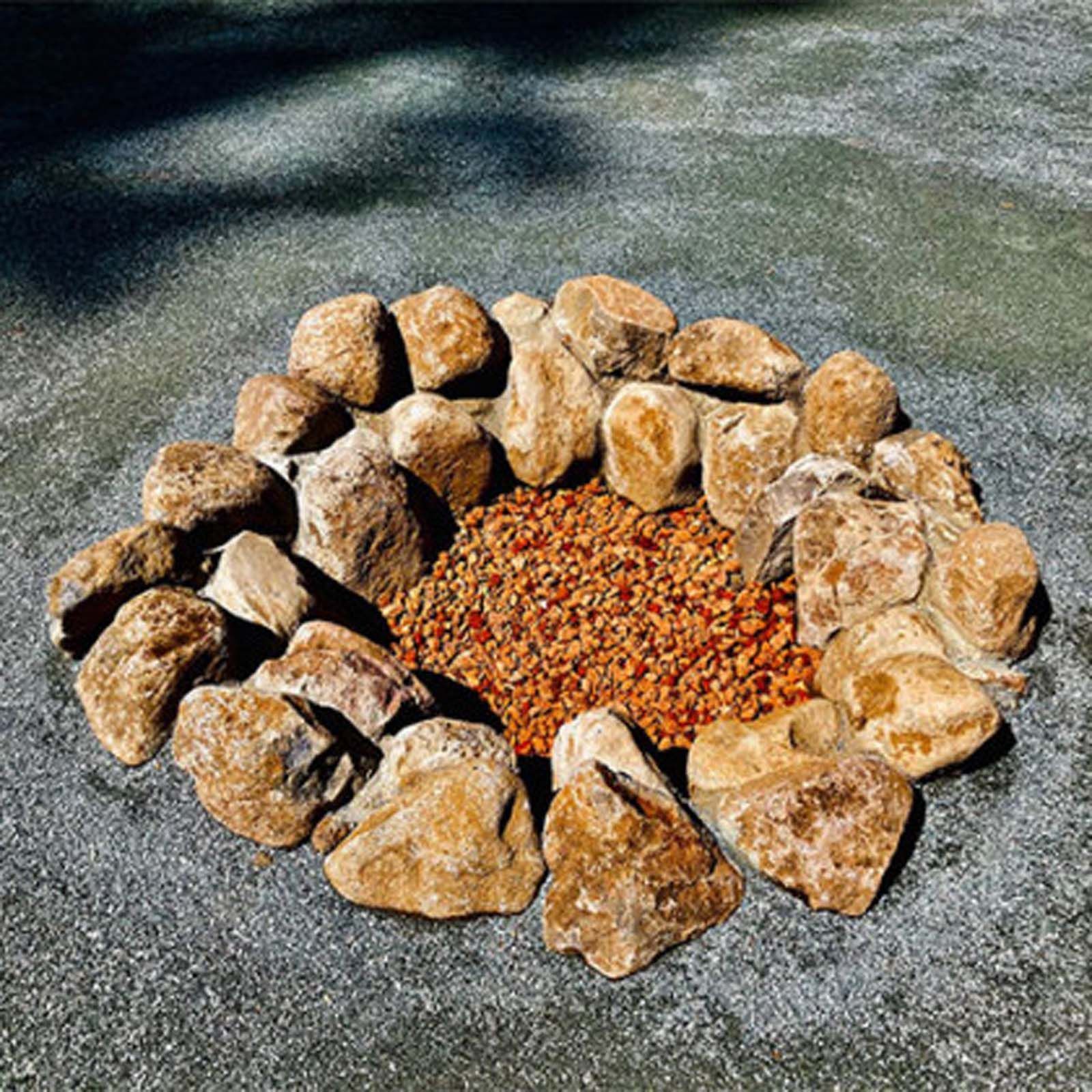 A circular fire pit made of tan rocks filled with small, reddish-brown gravel, set on a gray, paved surface.