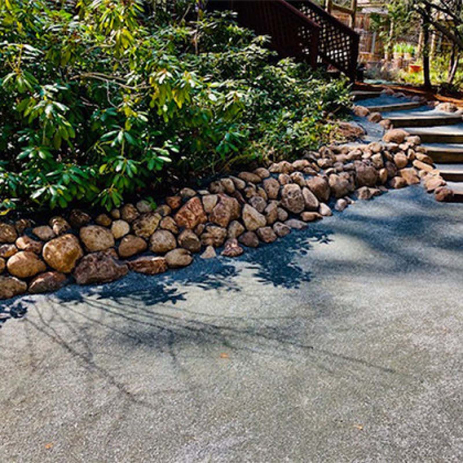 A rock retaining wall borders a paved walkway that leads up stone steps near a lush green garden.