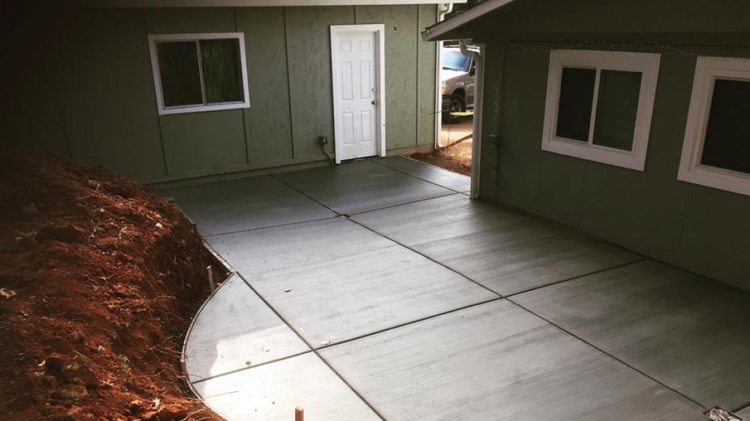 A newly poured concrete driveway sits between two sage green building sections, adjacent to a sloped dirt embankment.